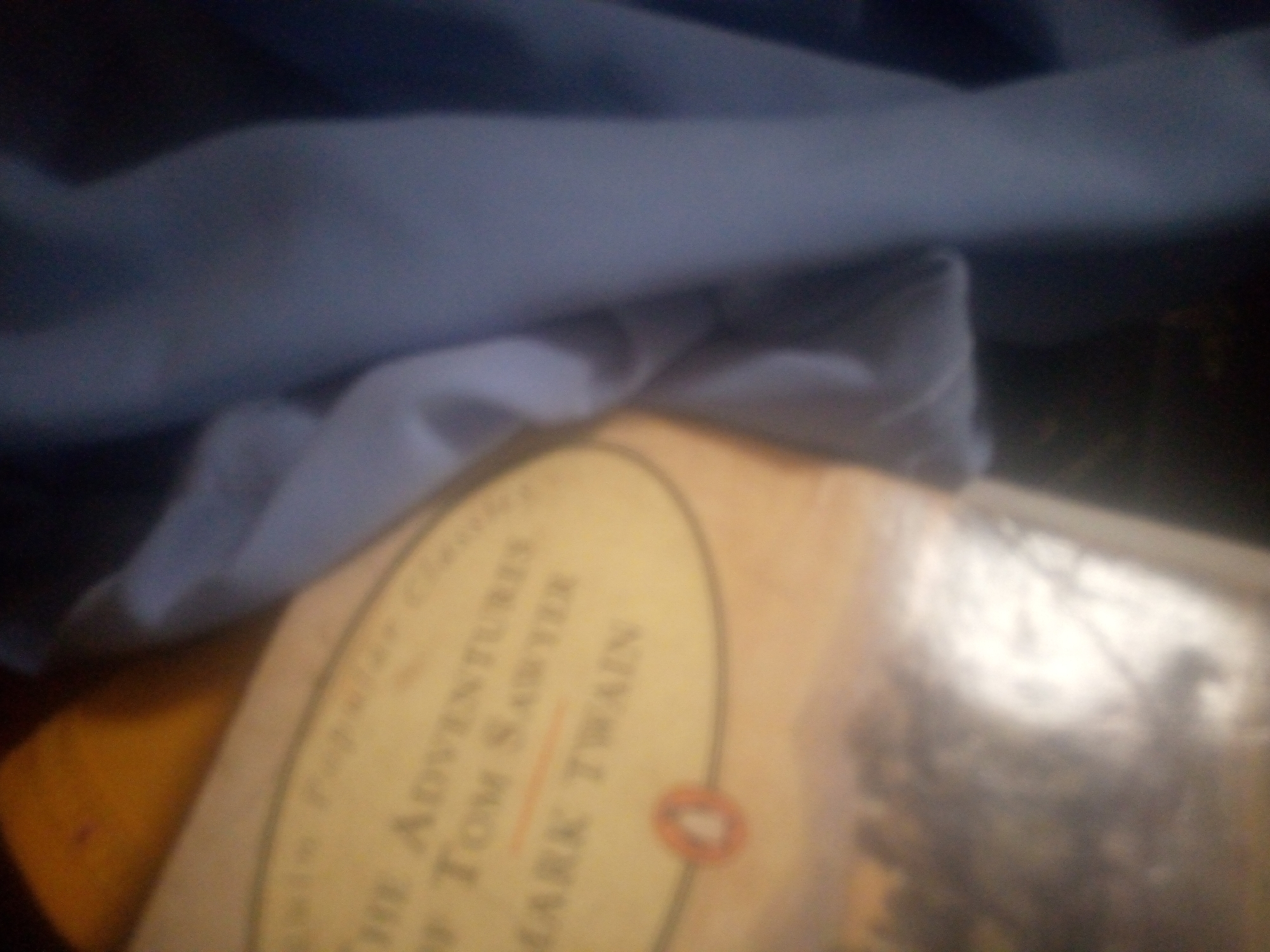 The image is a blurry, overhead shot of a book. The book appears to be an edition of "The Adventures of Tom Sawyer" by Mark Twain. The cover is a light yellow with black text and an oval logo. The book is lying on a blue cloth. The background is dark and out of focus. The image was taken indoors, likely in a home or library. The location is Unknown, Unknown.