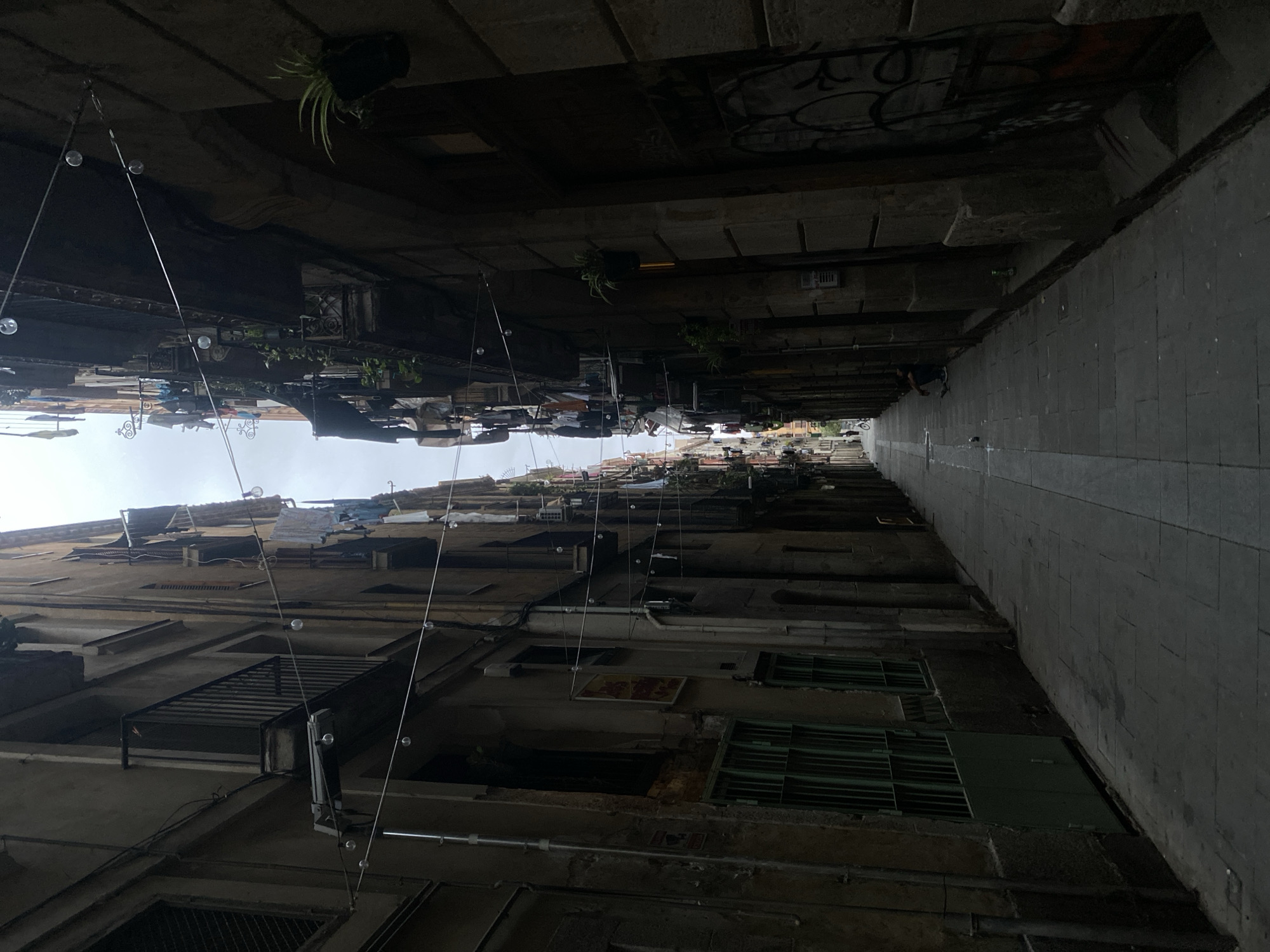 This image depicts a narrow pedestrian alleyway in Barcelona, Spain, likely within the Gothic Quarter or a similar historic district, characterized by its tall, aged buildings. The sky above is overcast, providing diffuse light that struggles to reach the street level.
The alley runs straight into the distance, flanked by multi-story stone and stucco buildings. On the left, buildings feature multiple windows, some with green metal security grates on lower levels, and several balconies with black iron railings. Vertical utility pipes are visible along the walls. A faded yellow and red sign is affixed to one building section, below a window.
On the right, similar aged buildings rise, many displaying clotheslines with various items of laundry hanging to dry from balconies and windows. Potted green plants are mounted on the walls and balconies. Near the foreground on the right, the lower section of a building wall is covered with black and white graffiti. Several strings of small, clear spherical lights are suspended across the alley between the opposing buildings, forming a crisscross pattern overhead.
The ground is paved with rectangular stone slabs, featuring a distinct lighter strip running centrally down the length of the alley. In the mid-distance on the right side of the alley, a single individual is visible, crouched or sitting on the ground near the wall, wearing dark clothing. Small pieces of debris are scattered on the pavement near this person. Further down the alley, a faint figure, possibly on a bicycle, can be seen. The general atmosphere is somewhat shadowed and enclosed due to the height and proximity of the buildings.