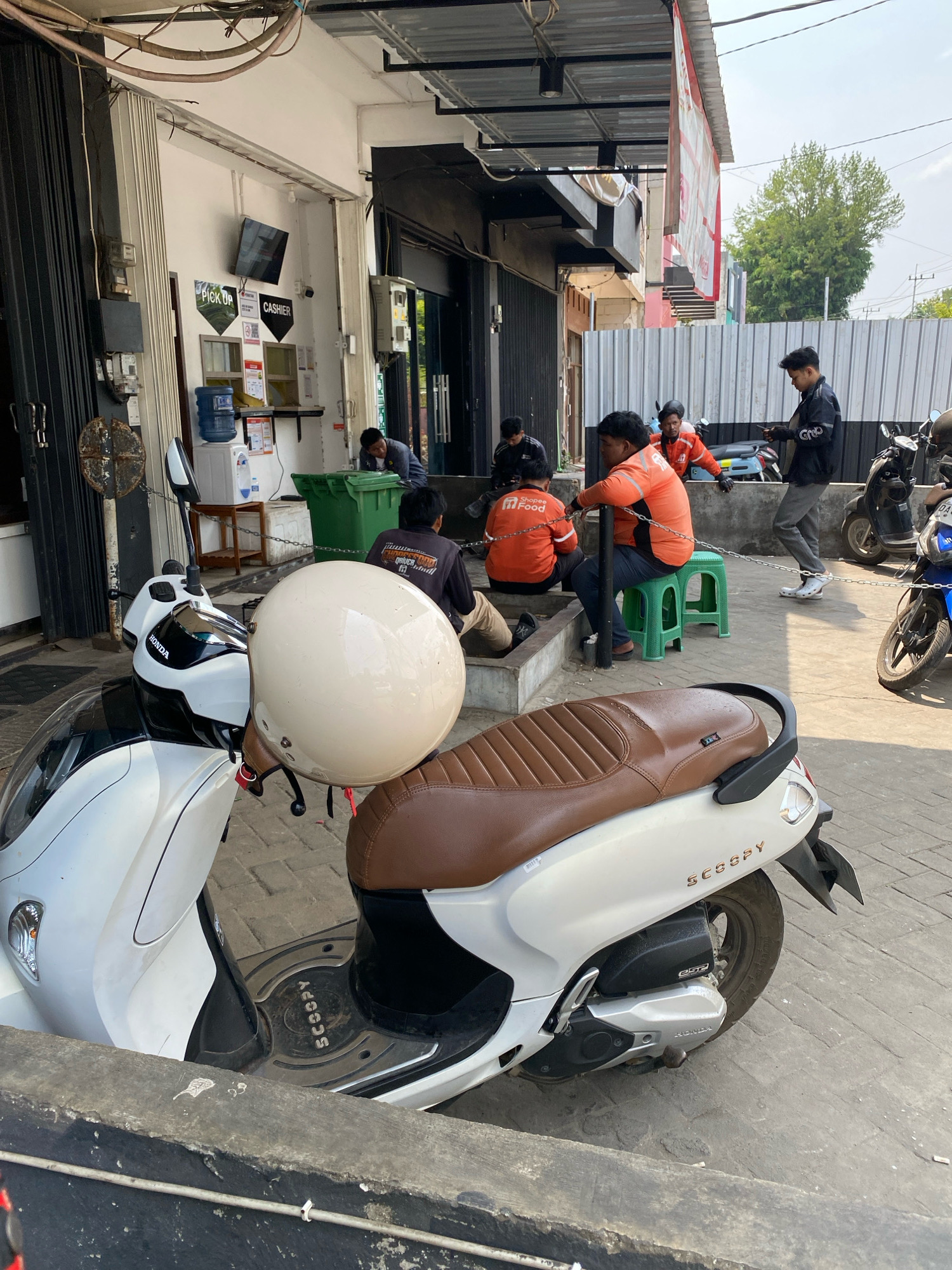 In the city of Banjarbaru, Indonesia, a group of people are gathered outside a building. A white Honda Scoopy scooter is parked in the foreground, with a cream-colored helmet resting on its handlebars. The building appears to be a food establishment, with signage indicating "Pick Up" and "Cashier." Several individuals, some wearing orange vests with "Shopee Food" logos, are seated on a green plastic stool or on the ground. One person in a black jacket is standing and looking at their phone. The environment is outdoors, with a paved ground surface and a metal fence in the background. The lighting suggests a bright, sunny day.