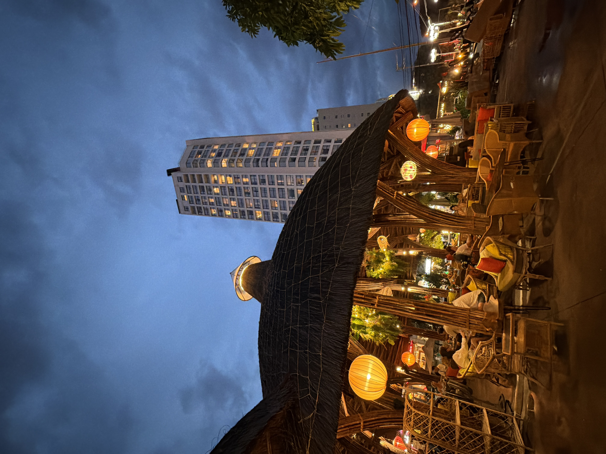 The image depicts an outdoor seating area of a restaurant or cafe in Nha Trang, Vietnam, at dusk. The foreground is dominated by a thatched-roof structure with bamboo supports, illuminated by warm-toned paper lanterns and string lights. Several patrons are seated at tables and chairs, engaged in dining or conversation. In the background, a tall, modern high-rise building stands out against the twilight sky, its windows illuminated, indicating it's likely residential or an office building. The overall atmosphere is cozy and inviting, with the natural materials of the outdoor structure contrasting with the urban backdrop.