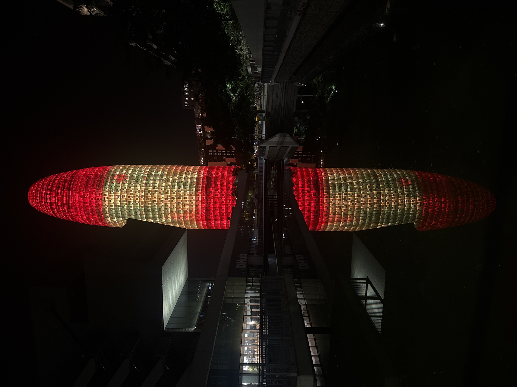 The image shows the Agbar Tower (Torre Glòries) at night in Barcelona, Spain. The tower is illuminated with red and yellow lights, creating a vibrant contrast against the dark sky. A body of water in the foreground reflects the illuminated tower and the surrounding architecture. On the left, a modern building with a glass facade and a cantilevered section is visible. The reflection in the water shows the tower's illumination and a portion of the building's structure. The scene appears to be in a public urban space, possibly a plaza or promenade, with trees and pathways visible in the background. The overall atmosphere is one of urban dynamism and architectural spectacle at night.