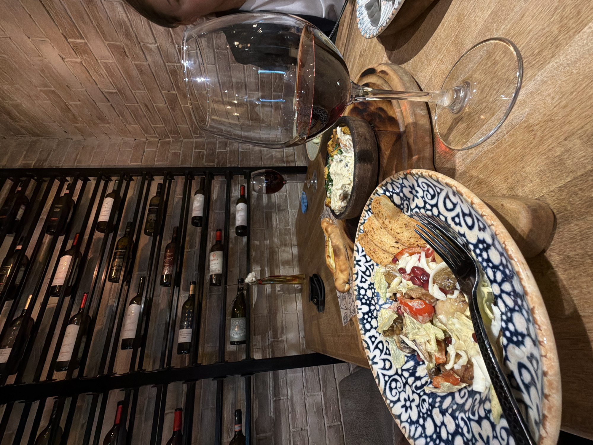 The image shows a table setting in what appears to be a restaurant, possibly located in Izhevsk, Russia, given the context. A large wine glass filled with red wine dominates the upper portion of the image, partially obscuring an individual at the very top edge. Two bowls containing food are visible. One contains a creamy dish with greens, the other a salad with toast. A fork rests on the salad. The table is made of wood. Behind the table and to the left, a wine rack displays an assortment of wine bottles mounted on the wall. The wall behind the rack is textured with what appears to be brick.