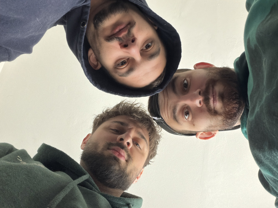 The image displays three adult males looking down directly into a camera positioned at a low angle. The background is an undifferentiated, plain light-colored surface, consistent with an interior wall or ceiling.
To the left, one male wears a dark green hooded garment, has light brown, medium-length hair, and a dark beard; his lips are slightly pursed. To the right, another male wears a dark blue hooded sweatshirt with the hood up, possesses dark hair, and a beard and mustache; his lips are also slightly pursed. Centrally positioned and slightly lower, the third male wears a dark green garment, a dark baseball cap worn backward, and a full beard; his eyes are wide open, directed at the lens. A mole is visible on his left cheek.
The scene is evenly illuminated. The image provides no discernible visual cues to pinpoint a specific location within the city of Varna, Bulgaria; however, the background suggests an interior setting.