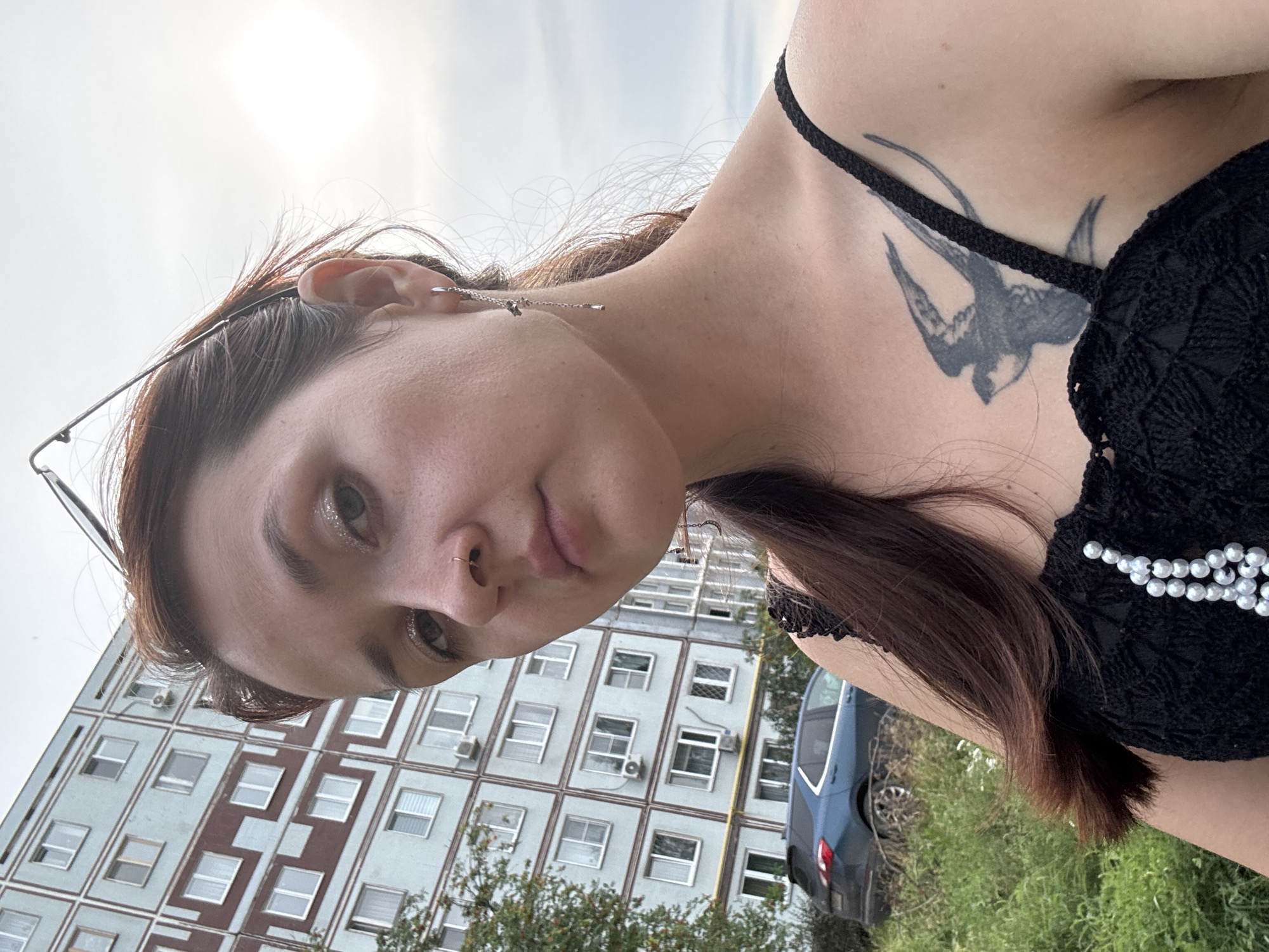 A woman with brown hair and a tattoo of a bird on her chest is taking a selfie outdoors. She is wearing a black top with beaded embellishments and a silver earring. Behind her, a light blue apartment building with maroon trim is visible, characteristic of the architecture found in Naberezhnye Chelny, Russia. A blue car is parked on the street in front of the building, surrounded by green grass and trees. The sky is overcast, suggesting a cloudy day.