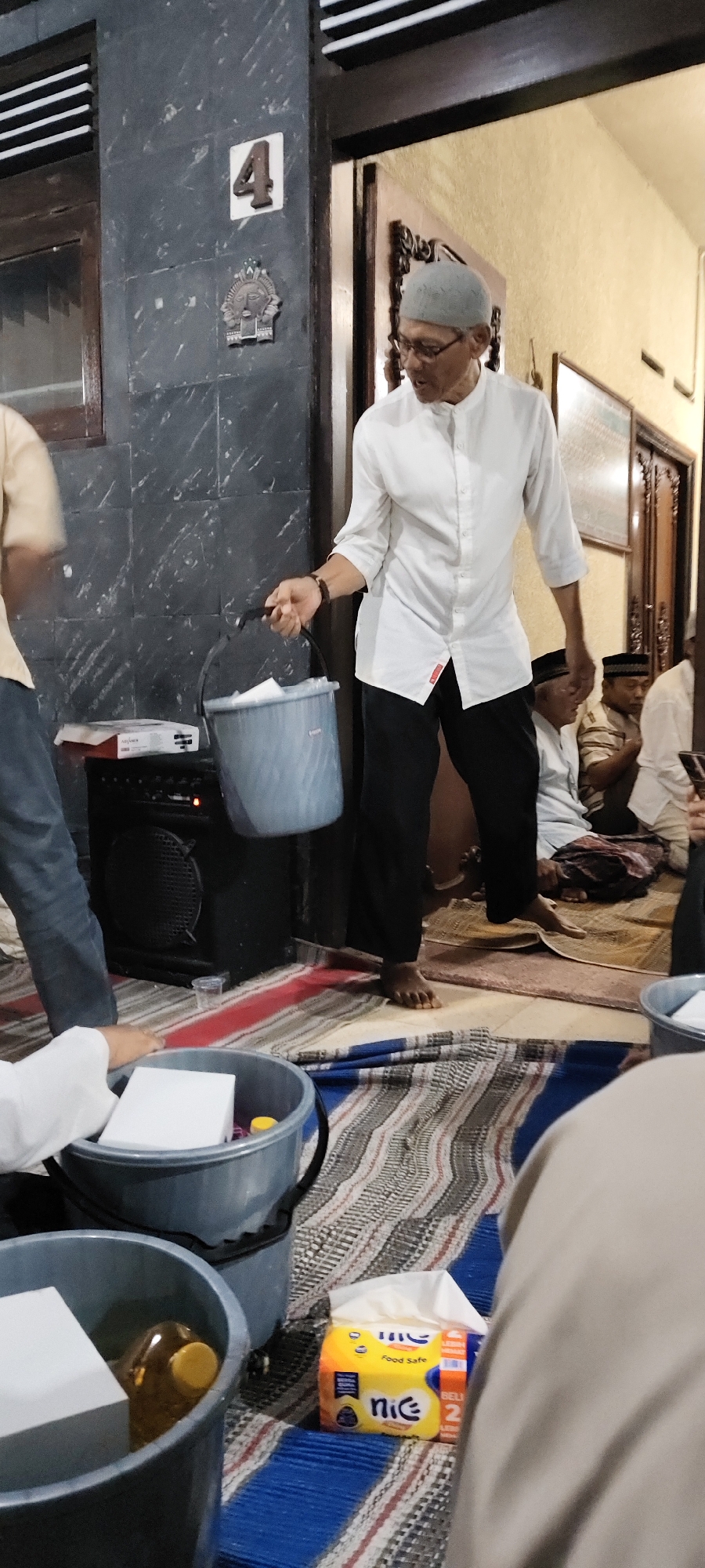 A man in a white shirt and grey cap holds a bucket filled with white items. He is standing in a room with patterned mats on the floor. A speaker is visible behind him, and a number '4' is displayed on a wall. In the foreground, two more buckets are present, one containing white objects and a bottle of yellow liquid, and another with white items and a pack of "nic" brand tissues. Several other people, dressed in traditional attire, are seated on the mats in the background. The scene suggests a gathering or event taking place indoors, possibly in Malang, Indonesia, given the cultural context implied by the attire and possibly the architecture.