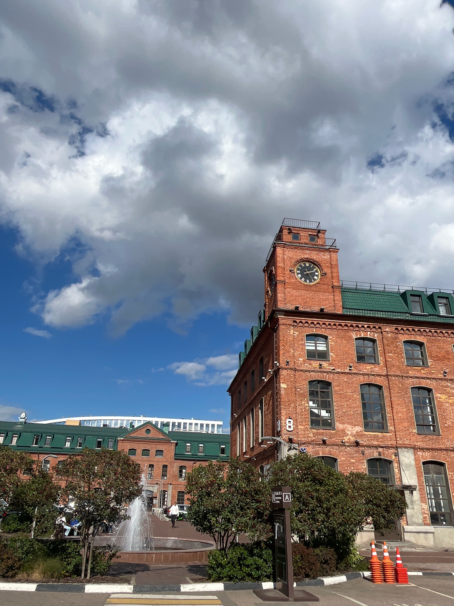 The image shows a brick building with a clock tower. The building is red brick with a green roof. The clock tower has a round clock face with black hands. There are windows on the building. In front of the building, there is a fountain with water spraying. There are trees and bushes around the fountain. In the background, there are other buildings and a blue sky with clouds. The location is in Moscow, Russia, at the Red October Chocolate Factory complex.