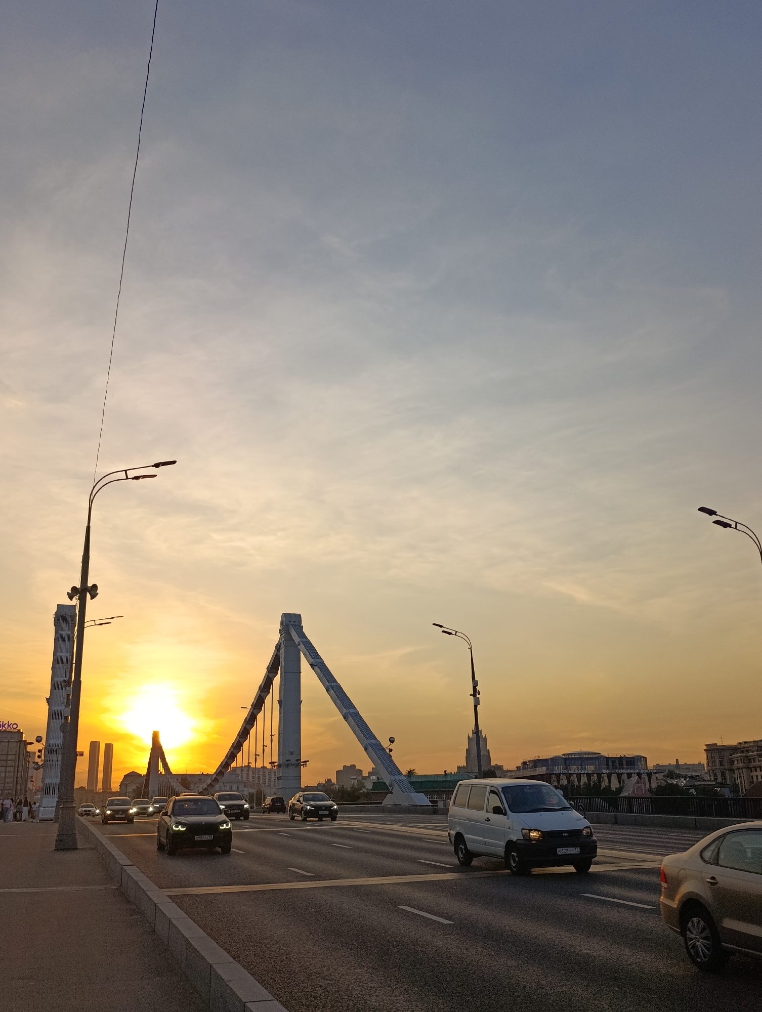 This forensic description details an urban scene captured during golden hour, likely sunset, on the Krymsky Bridge in Moscow, Russia. The sky, dominating the upper portion, transitions from a pale blue-grey at the zenith to a vibrant orange and yellow hue near the horizon, where the sun is visible as a bright, low-lying orb. Wispy, scattered clouds are present throughout the sky.
The prominent structure is the Krymsky Bridge, characterized by its distinctive white, A-frame cable-stayed pylons, from which numerous tension cables descend to support the bridge deck. The bridge deck accommodates a multi-lane roadway, with at least three lanes visible in the direction of the viewer.
Several vehicles are in motion on the bridge. In the foreground-center, a dark-colored BMW SUV with its headlights illuminated travels towards the viewer. To its right, a white Toyota Noah minivan is in an adjacent lane, also with headlights on. Partially visible on the far right is the rear of a gold-colored sedan, likely a Volkswagen Polo. Several other cars are visible in the distance, heading towards the sun, while others are seen traveling in the opposite direction. Road markings, including white lane dividers and a yellow curb line, are discernible on the asphalt.
On the left side of the image, a concrete sidewalk runs parallel to the roadway, bordered by a raised curb. Multiple pedestrians are visible walking on this sidewalk. A tall streetlamp with two curved arms and luminaires stands on the sidewalk, accompanied by a utility pole featuring what appear to be speakers or cameras. Another similar streetlamp is positioned further down the road on the right.
In the background, various urban buildings are silhouetted against the sunset glow. On the far left, a building with partial text "ikko" is visible. Further in the background, beyond the bridge, a building features Cyrillic script identifying it as a "МЕДИЦИНСКИЙ ЦЕНТР" (Medical Center) and "СЕМЕЙНЫЙ ДОКТОР" (Family Doctor). A thin, dark utility cable descends diagonally from the upper-left corner of the frame, traversing the sky.