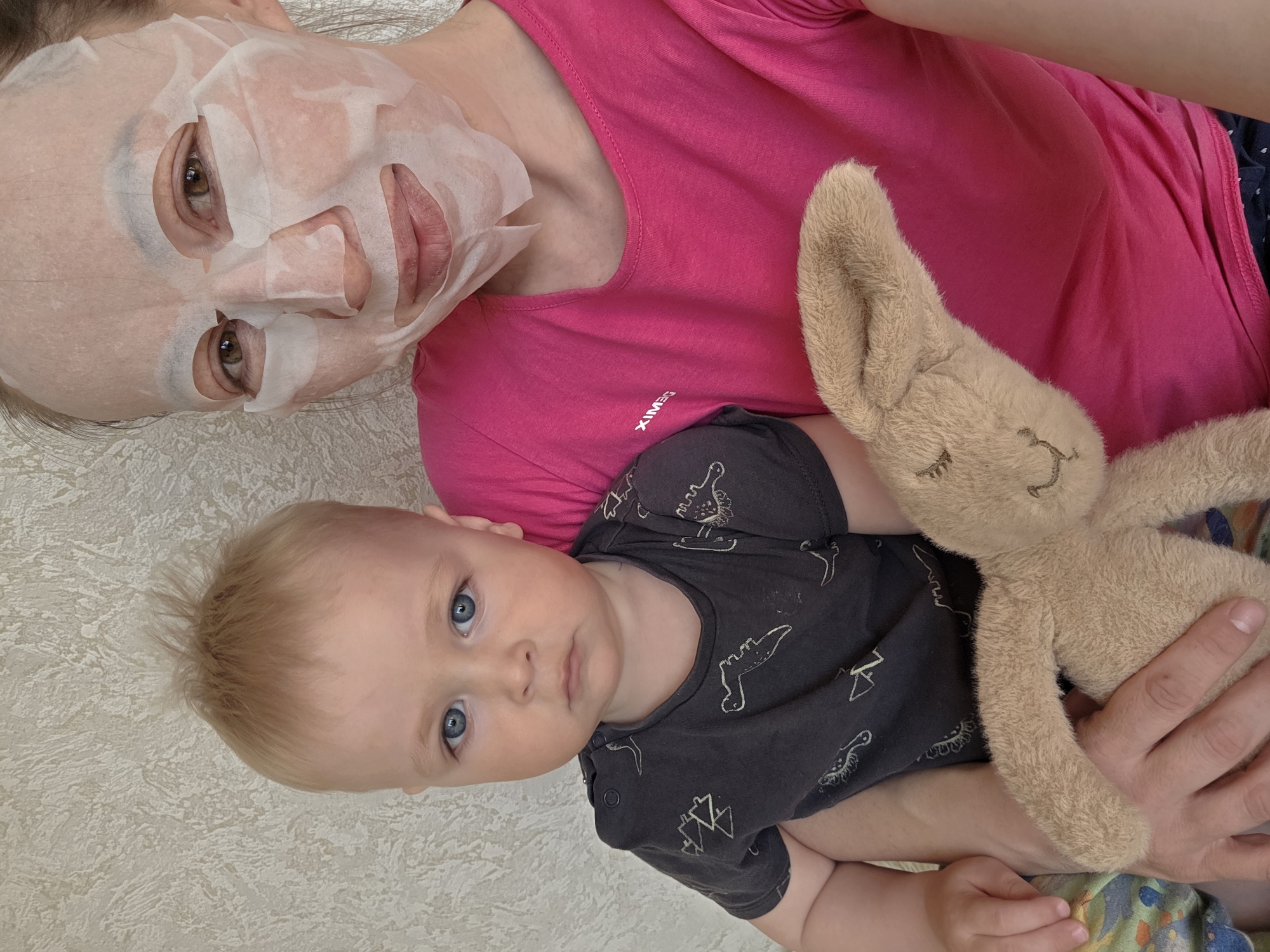 An overhead shot captures a woman and a baby lying down together in Chaikovsky, Russia. The woman, positioned at the top left, is wearing a white sheet face mask, revealing her brown eyes and parts of her lips. She has light brown hair and is wearing a bright pink t-shirt. The word "XIMEG" is visible in white text on her shirt.
Beside her, in the lower right, lies a baby with fair skin, light blonde hair, and striking blue eyes, looking directly towards the camera with a calm expression. The baby is wearing a dark grey or black t-shirt adorned with light-colored patterns, possibly depicting dinosaurs or geometric shapes. The baby's small hands are visible near a cuddly, light brown stuffed bunny. The bunny, with closed eyes embroidered on its face, is held by either the baby or the woman.
The background features a light-colored, textured wall or surface. The overall scene suggests a relaxed, intimate moment between a parent and child, possibly during a quiet time at home.