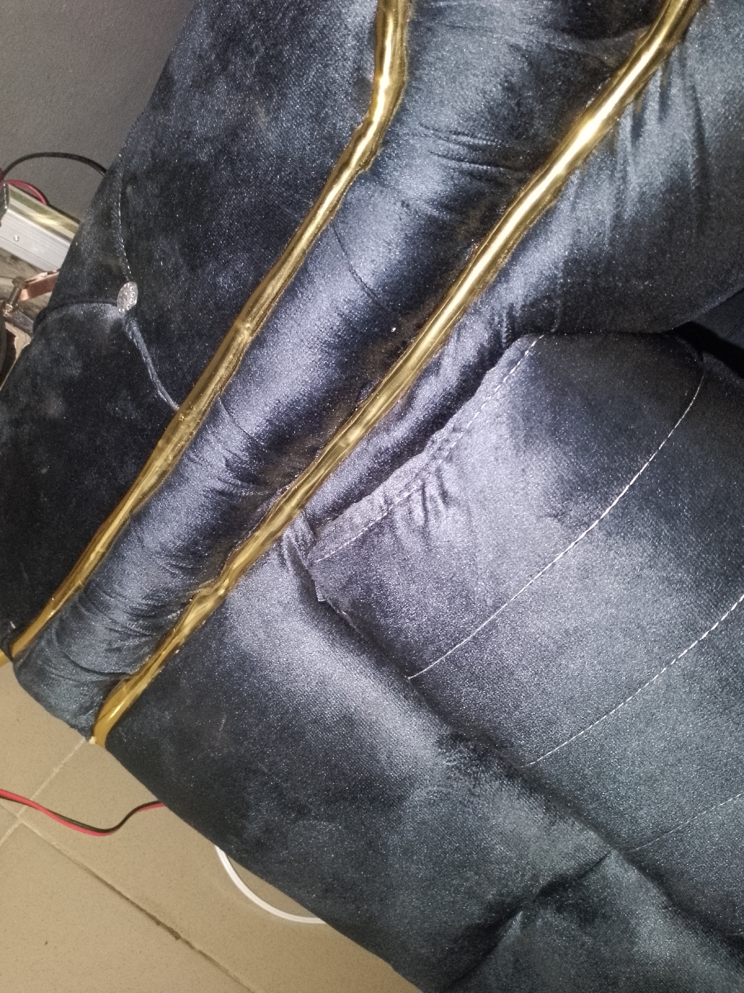 The image shows a close-up, overhead view of a dark blue, possibly velvet, upholstered surface with gold piping along its edges. The upholstery appears to be part of a piece of furniture, possibly a chair or sofa, with tufting visible in some areas and stitched details in others. A small, circular, crystal-like embellishment is visible on the left side of the frame. Some electronic components and wiring are also present in the upper left and lower left portions of the image. The surface is resting on a beige tiled floor. The scene appears to be indoors, in an unknown location within the city of Unknown, Unknown.