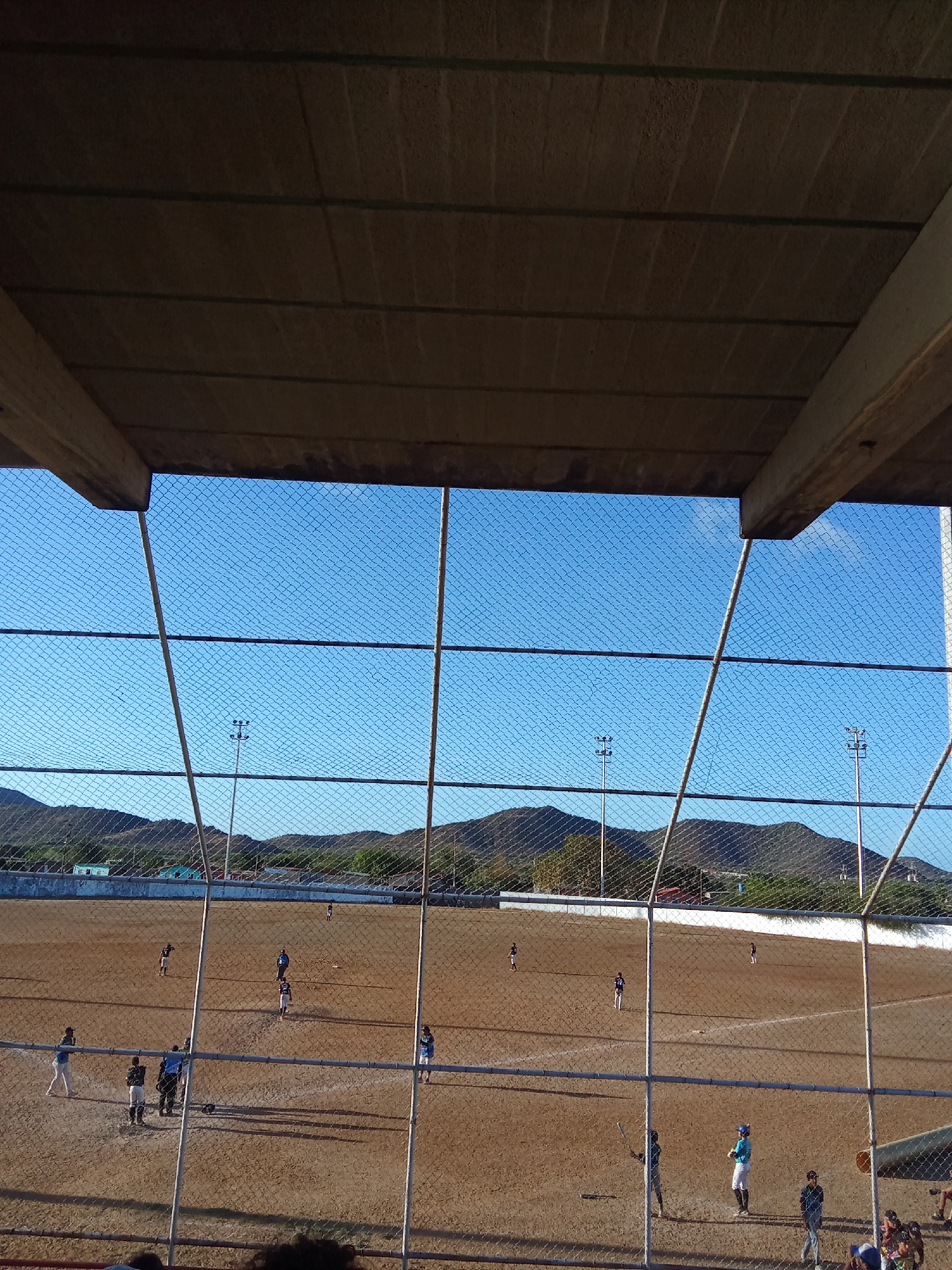 The image shows a baseball game in progress, viewed from behind a chain-link fence under the cover of a stadium structure. The playing field is mostly brown dirt, with white lines marking the base paths. Mountains are visible in the background under a clear blue sky. Several players are scattered across the field, some near home plate and others positioned at different bases. Light poles stand along the perimeter of the field. The location is Juan Griego, Venezuela.
