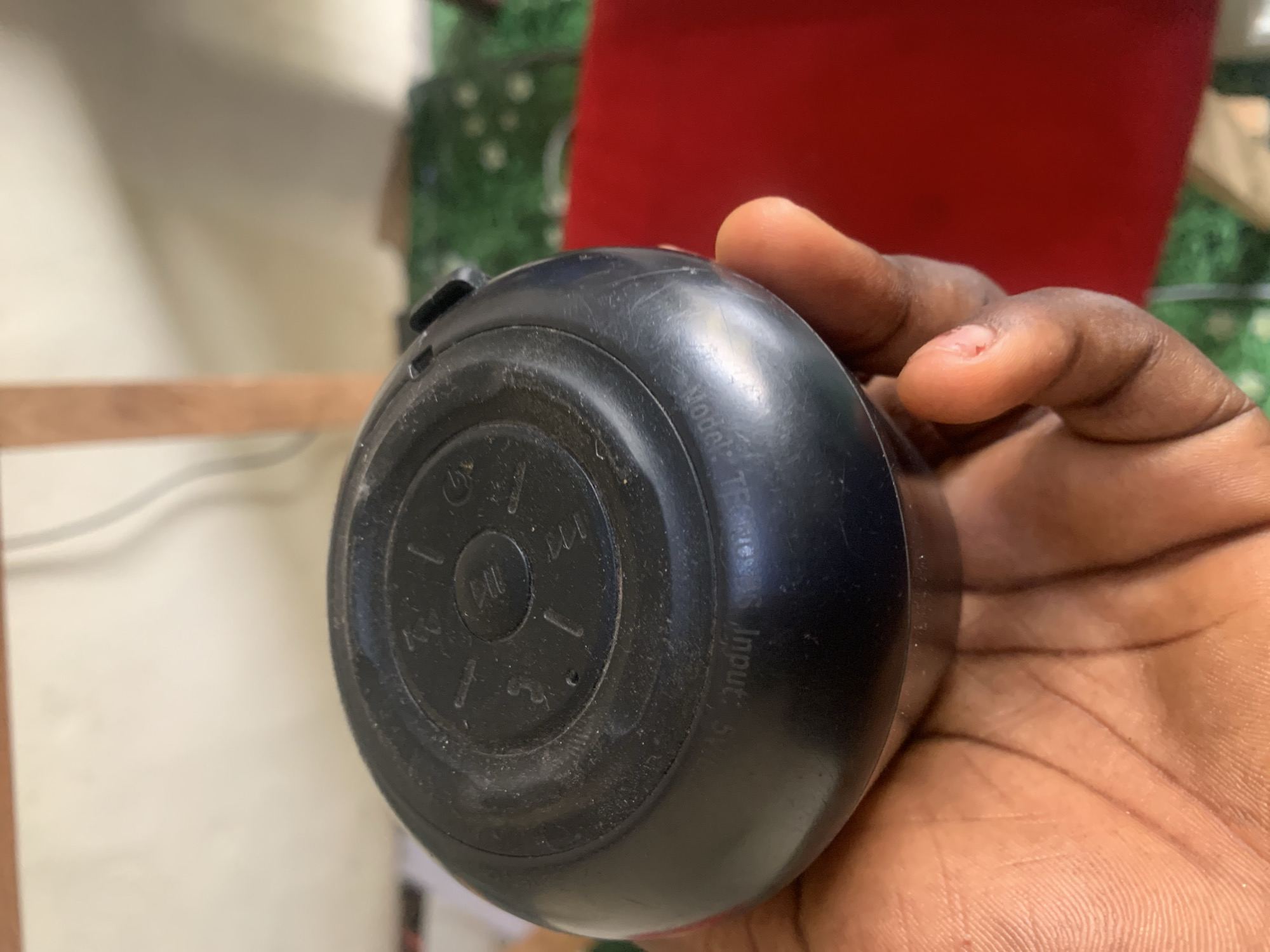 In Jauro Yinu, Nigeria, a close-up shot captures a person's hand holding a black, round electronic device. The device appears to be a speaker, with embossed text on its underside reading "Model: TFC-808S Input: 5V". Some dust is visible on the surface of the device. The background is blurred, showing hints of a light-colored wall, wooden structures, and green foliage, suggesting an indoor or outdoor setting within a residential area. The lighting is natural, casting soft shadows.