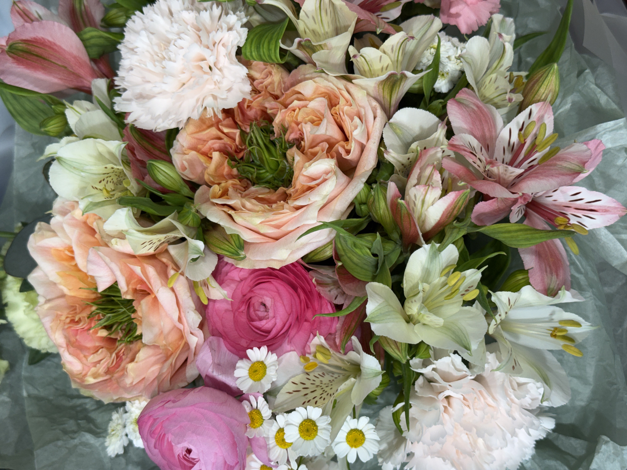 A close-up, overhead shot showcases a vibrant floral arrangement. The bouquet is dominated by large, ruffled blooms in shades of peach, pink, and cream. Among these are smaller white flowers with yellow centers, resembling daisies, and delicate white and pink alstroemeria. The arrangement is set against a textured, pale green background, likely wrapping paper. The lighting is soft and even, highlighting the intricate details and textures of the petals and foliage. No people or specific location are visible.