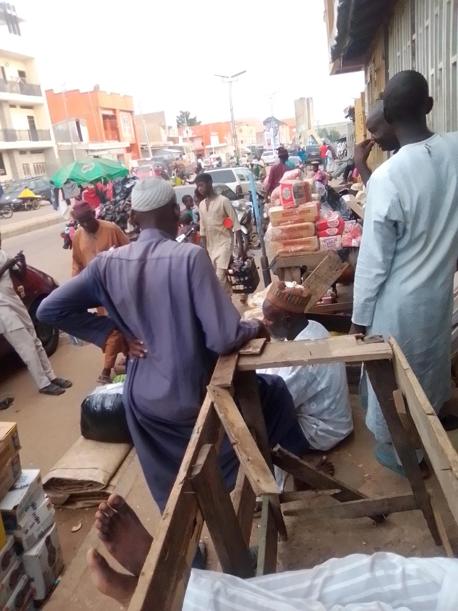 The image depicts a busy marketplace scene in Arawa, Nigeria. Multiple individuals are present, some engaged in transactions or simply standing around. In the foreground, a person wearing a purple tunic and a white cap is leaning on a wooden structure. To their right, another person in a white kaftan stands. A man in a traditional cap and white tunic is seated on the ground, looking down. In the background, various stalls are visible with goods stacked, including what appear to be loaves of bread packaged in red and white. A green umbrella shades a stall. Several vehicles, including a car and motorcycles, are parked on the street. The environment appears to be an urban street market with buildings lining the background. The overall impression is one of a lively commercial area with a mix of people and goods.