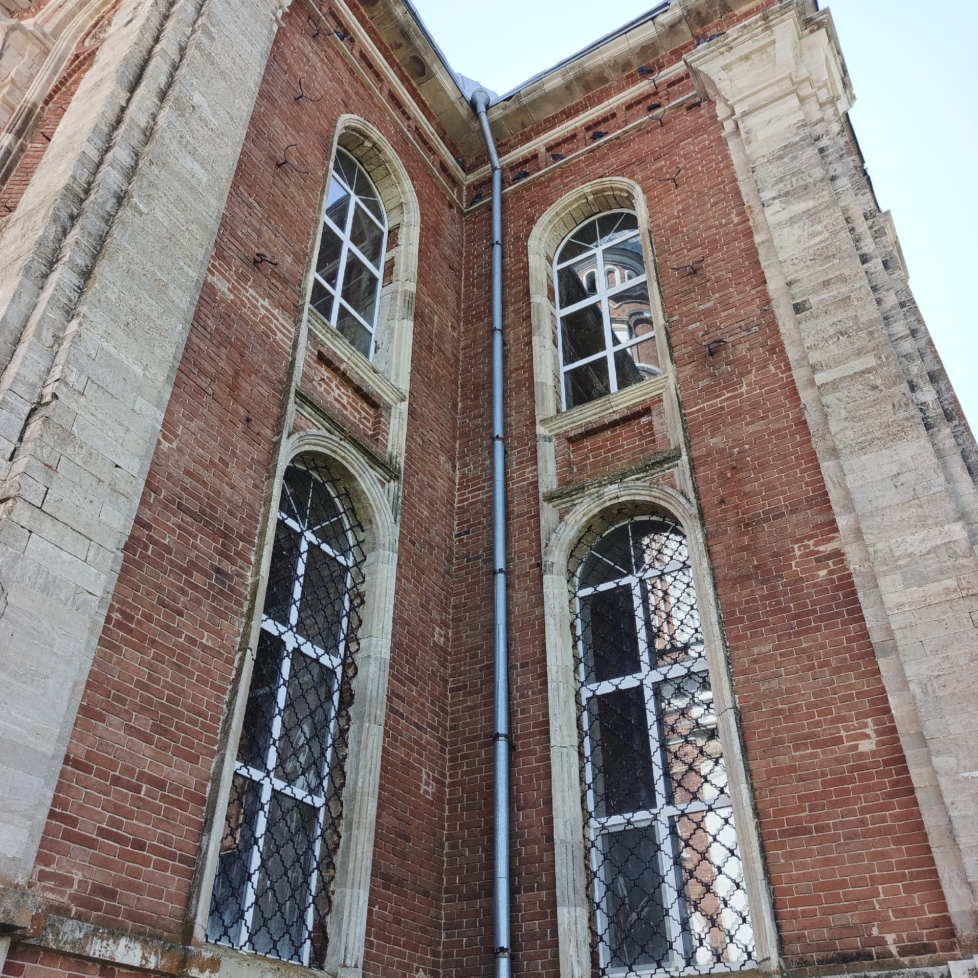 The image is a low-angle shot of the exterior corner of a red brick building, possibly a church or historical structure, under a clear sky. The building features large, arched windows with white frames and decorative metal grates. Some windows reflect an adjacent building with a dome. The corner of the building is accented with light-colored stone pilasters. A gray downspout runs vertically along the corner, attached with dark brackets. The brickwork shows signs of age and weathering, with some mortar missing. There are small, dark, hook-like objects embedded in the brickwork. The scene is devoid of people or any other discernible activity.