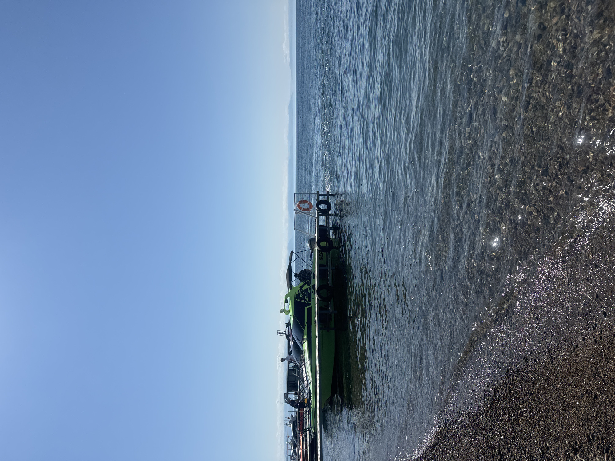 A bright green motorboat is docked at a wooden pier on a clear day in Listvyanka, Russia. The boat has a dark tinted windshield and a canopy providing shade. Two people are visible on the boat, one standing and one seated. The water is clear and shallow near the shore, with pebbles visible on the lakebed. The shoreline is covered in dark pebbles. In the distance, a hazy mountain range is visible on the horizon. A life preserver is hanging on the pier. Other boats are docked behind the green boat. The sky is a clear, bright blue with no clouds.