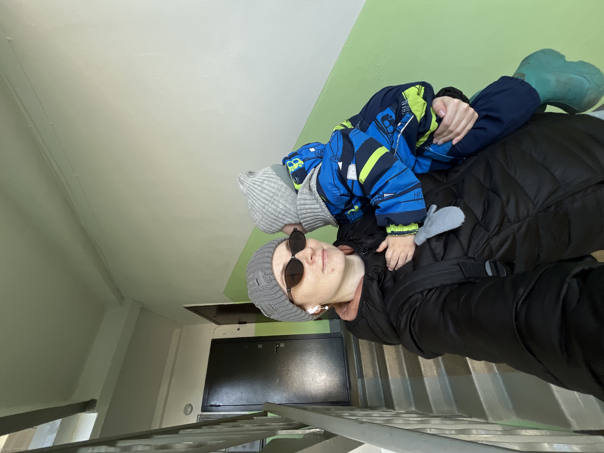 A woman wearing sunglasses and a gray beanie hat, carrying a child dressed in a blue and green jacket and gray hat, is positioned on a set of metal stairs. The woman is wearing a black puffy jacket, and the child has green rubber boots. The scene appears to be indoors, with light green and white walls visible behind them. A dark-colored utility box with a handle is mounted on the wall. Based on the photograph's context and metadata, the location is presumed to be in Chaikovsky, Russia. The subjects are likely posing for a selfie or casual photograph.
