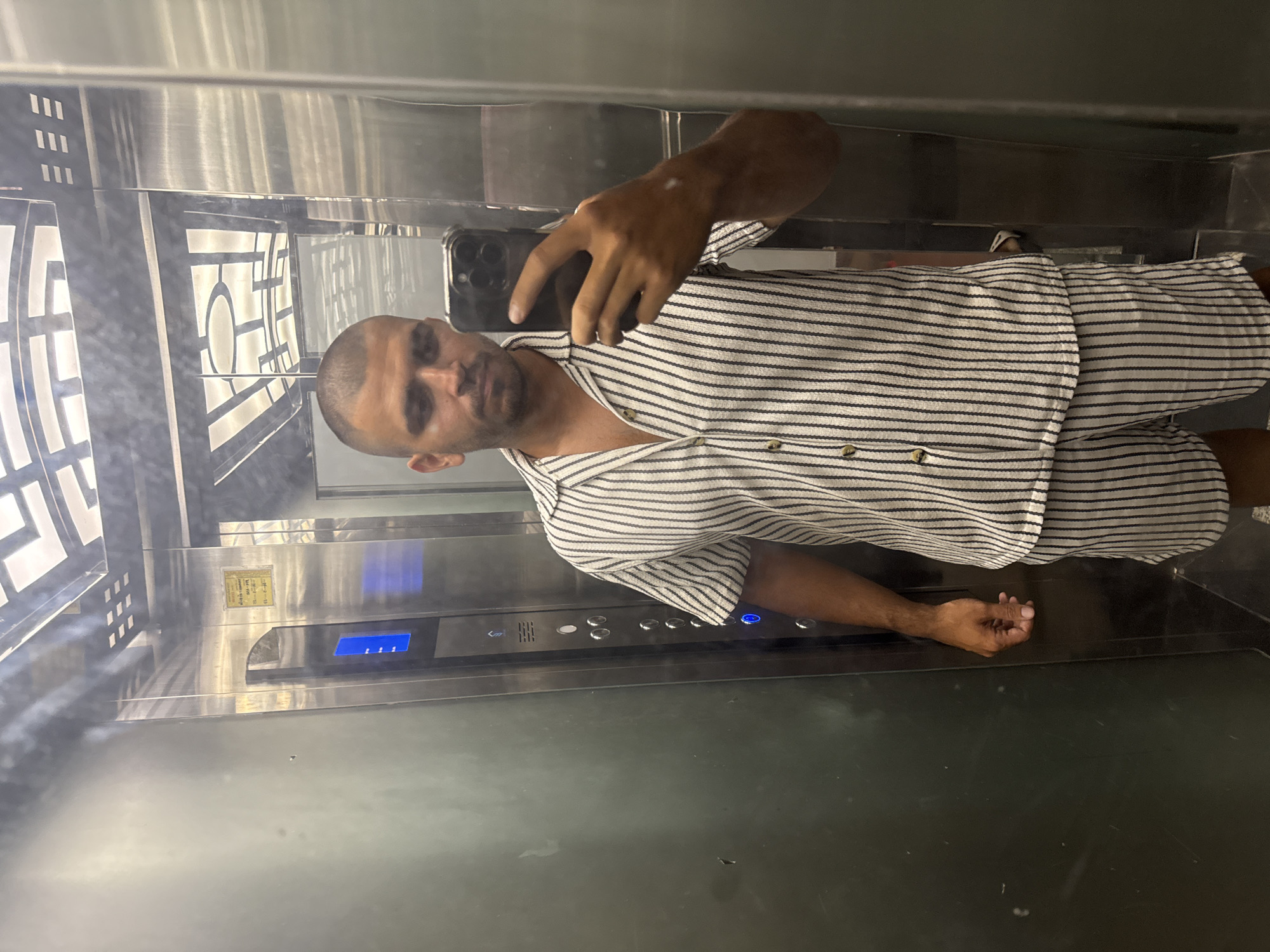 A person is standing in an elevator in a building in Đà Nẵng, Vietnam, taking a selfie in the mirror. The elevator has a metal interior, with a control panel with buttons. The person is wearing a striped short-sleeved button-down shirt and matching shorts. The elevator has decorative panels.