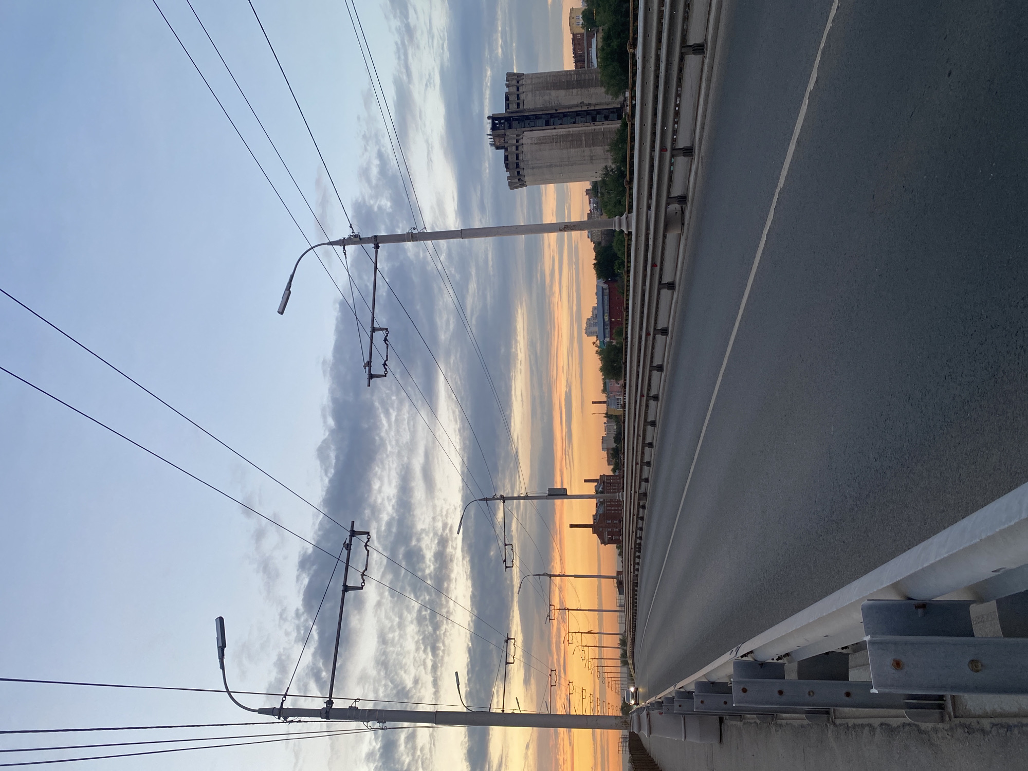 The image depicts a view of a road on a bridge in Novokuybyshevsk, Russia, during a sunset. The sky is painted with hues of orange, yellow, and blue, with scattered clouds. Overhead, a network of trolleybus wires crisscrosses the frame, supported by tall poles that also hold streetlights. The road itself is asphalt with a white dividing line and is lined with metal guardrails. In the distance, buildings and industrial structures are visible, including a prominent, tall, cylindrical concrete building. A single car is driving on the bridge, heading towards the vanishing point of the road. The scene is quiet, with no people visible on the bridge, suggesting it is either early morning or late evening.