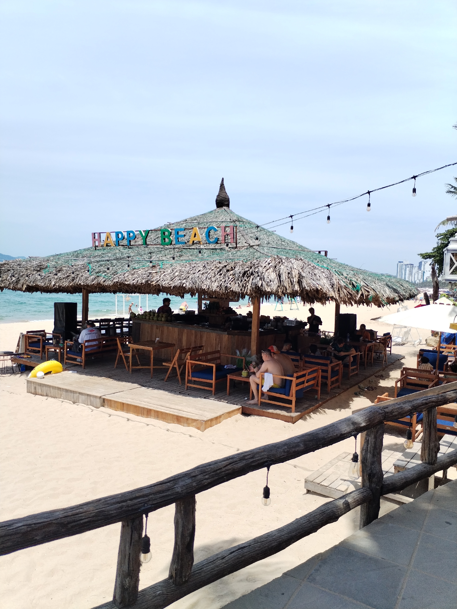 The image depicts a beach bar with a thatched roof, emblazoned with the words "HAPPY BEACH" in colorful letters, located in Nha Trang Ward, Vietnam. The bar is situated on a sandy beach, with the ocean visible in the background. Several people are seated at tables and chairs beneath the shade of the thatched roof, appearing to be patrons of the establishment. A wooden deck area surrounds the bar, with more seating and tables. In the foreground, a rustic wooden railing with hanging light bulbs overlooks the scene. The overall atmosphere is that of a relaxed beachside venue.