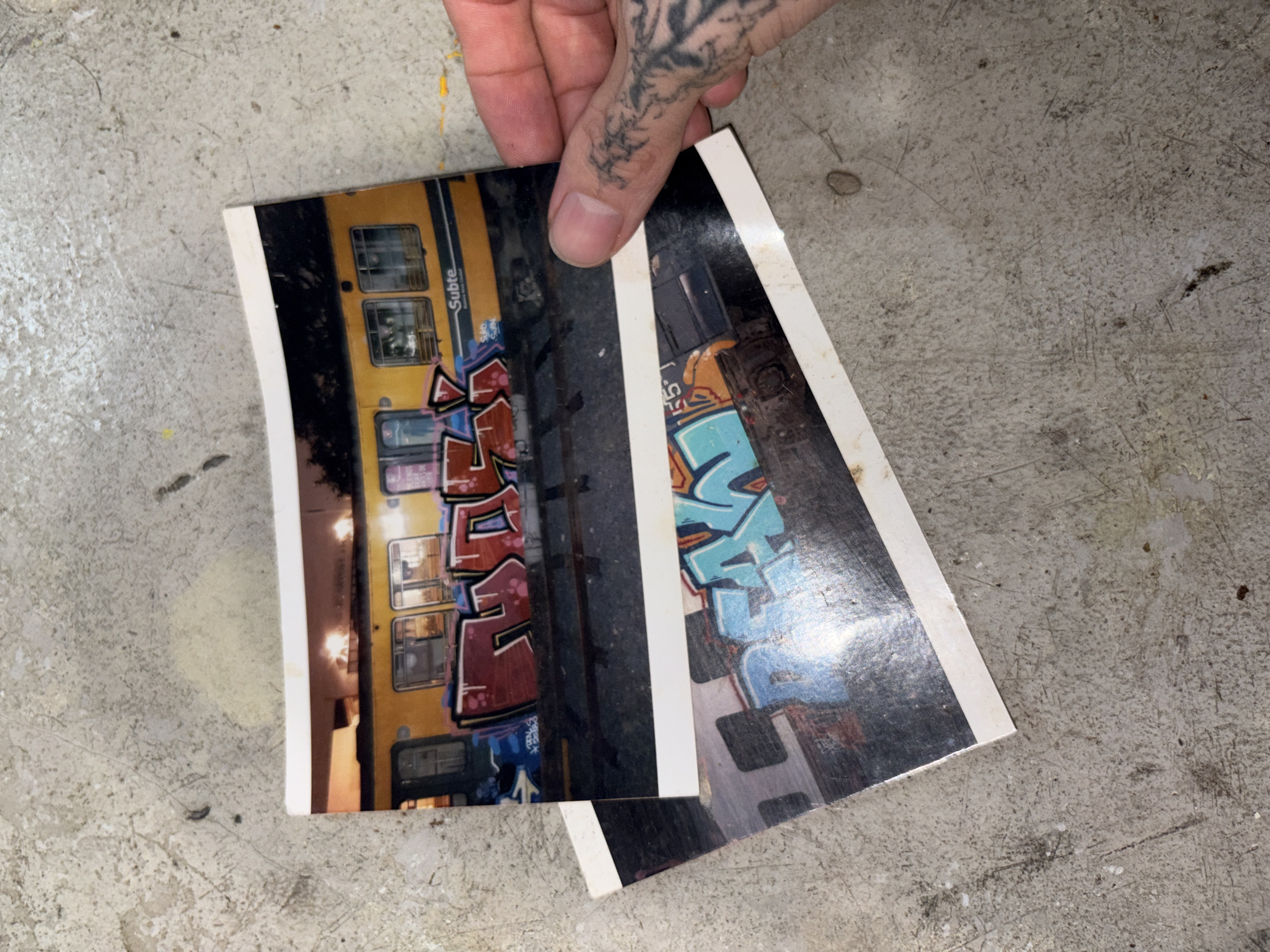 A person's tattooed hand holds two photographic prints, laid over each other, on a textured, light grey concrete surface. The hand, seen from above, displays a dark, intricate tattoo resembling branches or roots on the back of the thumb and extending towards the index finger.
The top photograph, predominantly visible, captures a yellow train car heavily covered in multi-colored graffiti. The graffiti features bold, stylized letters in shades of red, black, and white. Vertically written on the train's yellow body, above the graffiti and alongside train windows, the word "Subte" is clearly visible.
Partially obscured beneath the top print and the hand, the second photograph shows another section of graffiti, dominated by large, light blue letters with dark outlines. Both photographs have a white border. The concrete ground is rough, marked with scratches, scuffs, and small specks of debris. The scene implies an urban or industrial setting, possibly an outdoor area or a workshop. The "Subte" text on the train provides a strong location cue, indicating the subway system in Buenos Aires, Argentina.