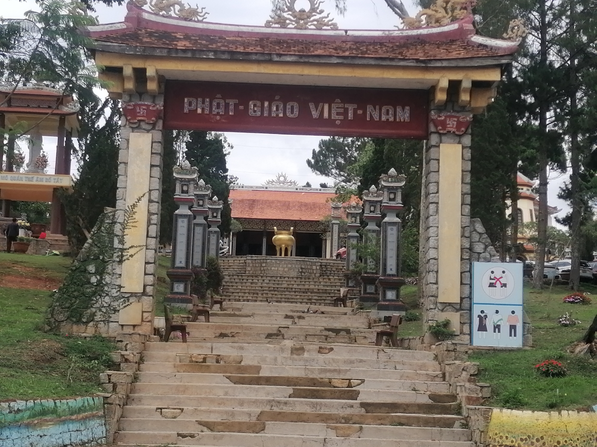 The image captures a view of a Buddhist temple in Da Lat, Vietnam. A stone archway, marked with the text "PHẬT GIÁO VIỆT-NAM" (Vietnamese Buddhism), frames a staircase leading towards the main temple building. The staircase is constructed of stone and shows signs of wear. A large, golden statue is visible in front of the temple. Additional structures and landscaping are present to the sides of the staircase. A sign depicting behavioral guidelines is located near the bottom of the stairs. The surrounding environment features trees and vegetation. A small building is visible to the left.