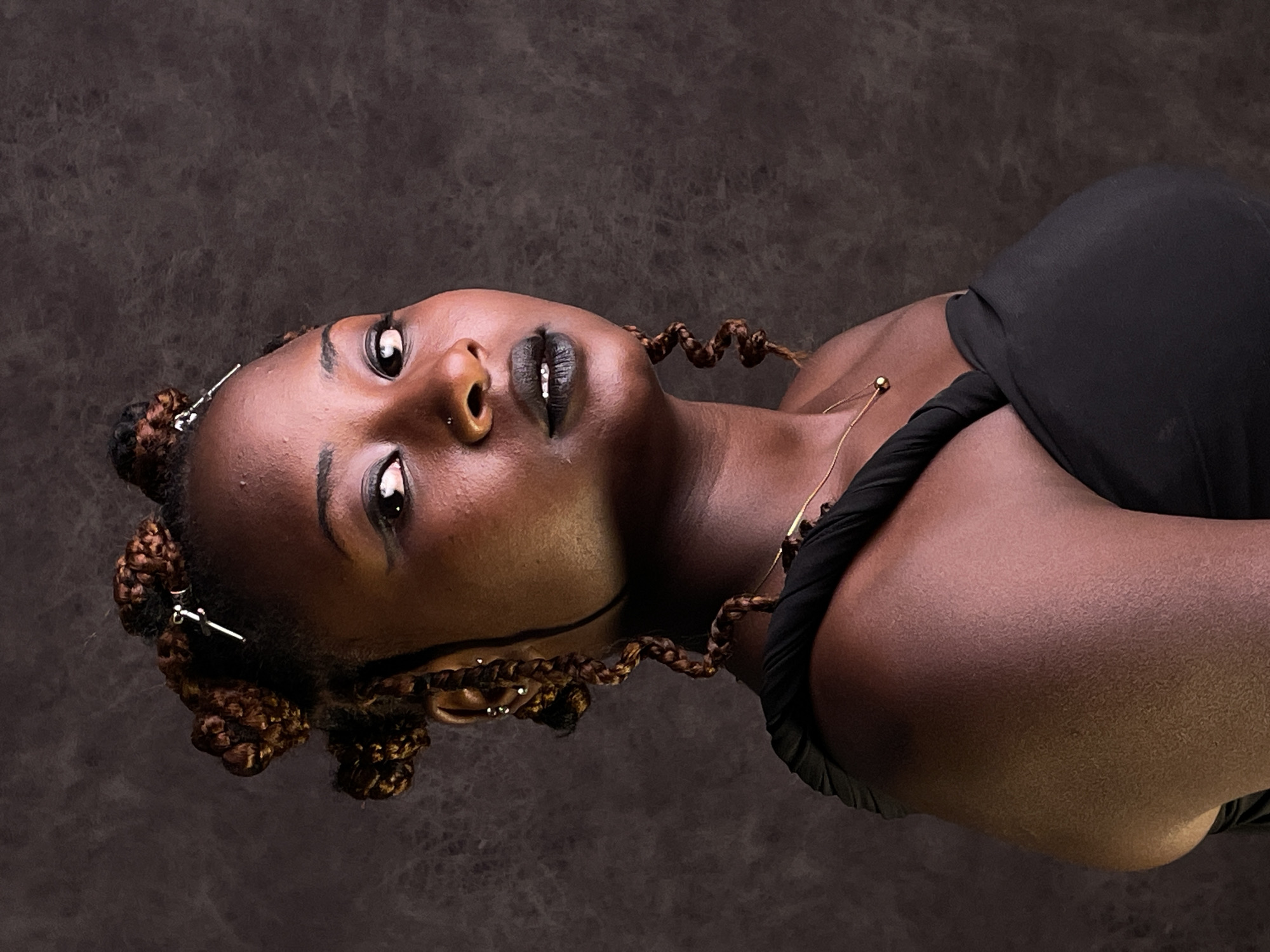 A close-up portrait captures a young Black woman with dark skin, her head tilted upwards and to the right against a dark, textured background. She gazes intently towards the upper left, conveying a contemplative or pensive emotion. Her features are accentuated by dramatic makeup, including thick black winged eyeliner and black lipstick with a small, sparkling detail on her lower lip.
Her hair is styled in elaborate brown braids, some coiled into small buns on the sides of her head and adorned with slender silver hair clips. Other braids frame her face and trail towards her shoulder. She wears a minimalist gold chain necklace with a small gold bead on her collarbone. Piercings include a silver stud in her right nostril and another above her upper lip. She is dressed in a simple black garment, possibly a top or dress, with some fabric twisted around her shoulder.
The lighting is soft, highlighting the contours of her face and the texture of her skin. The dark, uniform background suggests a studio setting, providing no specific outdoor location cues for Barakin Akawo, Nigeria. There is no visible text in the image.