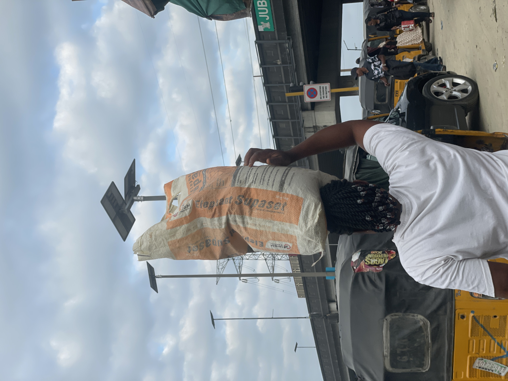 In Lekki, Nigeria, a woman carries a large, orange and white striped sack of what appears to be cement on her head, walking through a street scene with multiple auto rickshaws and a black SUV. She is wearing a white t-shirt and her braided hair, adorned with beads, is visible. In the background, an elevated highway with solar-powered streetlights stretches across the sky, with the word "JUBL" visible on a green sign. The sky is overcast with clouds. A "No Stopping, No Parking, No Waiting" sign is mounted on a yellow pole. Other pedestrians, some carrying bags, are seen in the distance, suggesting a busy market or transportation hub.