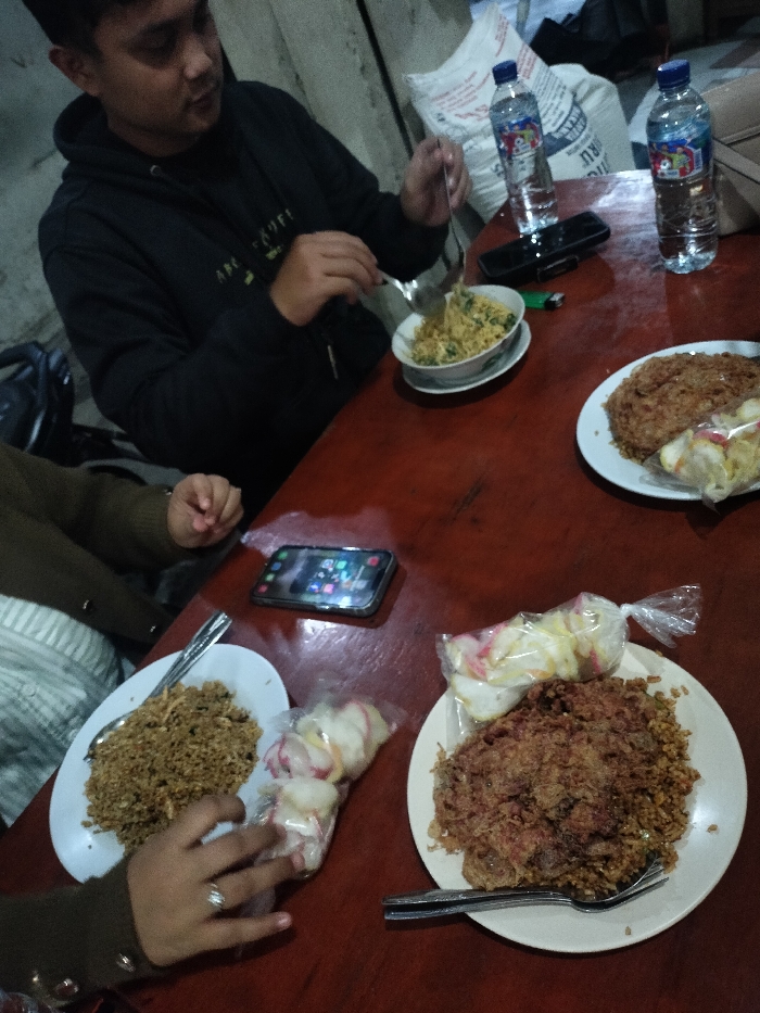 The image captures a casual meal shared by at least two people at a reddish-brown wooden table. A man, wearing a dark hoodie, is seated on the right side of the frame, actively eating fried rice from a small white bowl with a spoon and fork. A second person is partially visible on the left, with their hand near a plate of fried rice and a bag of crackers.
On the table are three plates of fried rice. The plate in the bottom right features a large, flat, crispy brown topping, resembling an omelet or shredded meat, alongside a plastic bag of colorful crackers (kerupuk). Another plate of fried rice is visible on the left, also accompanied by crackers in a bag. Two bottles of "AQUA" branded mineral water are present on the table, along with two smartphones (one displaying app icons) and a green lighter. In the background, behind the man, several large white sacks are visible.
The scene depicts individuals enjoying a meal, likely nasi goreng, which is a common dish in Indonesia. The dim lighting suggests it is either nighttime or an indoor setting with artificial illumination. The food and bottled water brand are consistent with the provided location context of Dramaga, Indonesia. The atmosphere appears relaxed, focused on the shared meal. Text on the "AQUA" bottles is legible, while text on the man's hoodie and the background sacks is not clearly discernible.