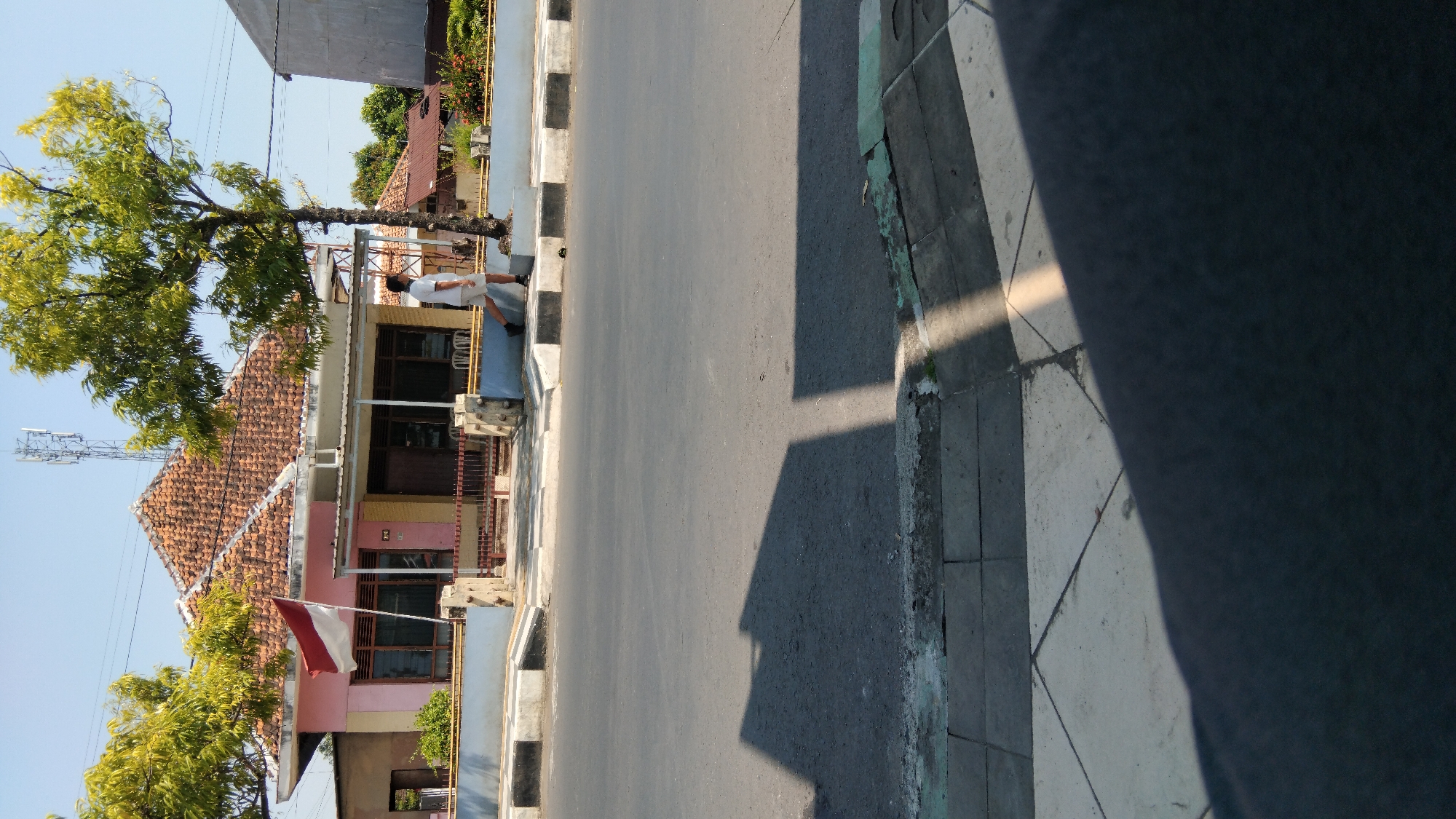 A medium shot captures a street scene in Batang, Indonesia, under a clear blue sky. The foreground is dominated by a gray asphalt road, with a sidewalk and a curb in the lower part of the frame. Sunlight casts distinct shadows across the road and sidewalk, indicating a bright, sunny day.
In the middle ground, a pink and yellow house with a tiled roof is visible. A red and white Indonesian flag flies from a pole in front of the house. To the right of the house stands a slender tree with green foliage. A person wearing a white t-shirt and light-colored shorts is walking on the sidewalk in front of the house, near the tree. The person is facing to the left and appears to be walking away from the camera.
The background shows more of the house, with awnings over the windows and doors. Beyond the house, other buildings and trees are visible, with a telecommunications tower rising above the rooftops. The overall impression is a snapshot of a typical residential street in a sunny climate.