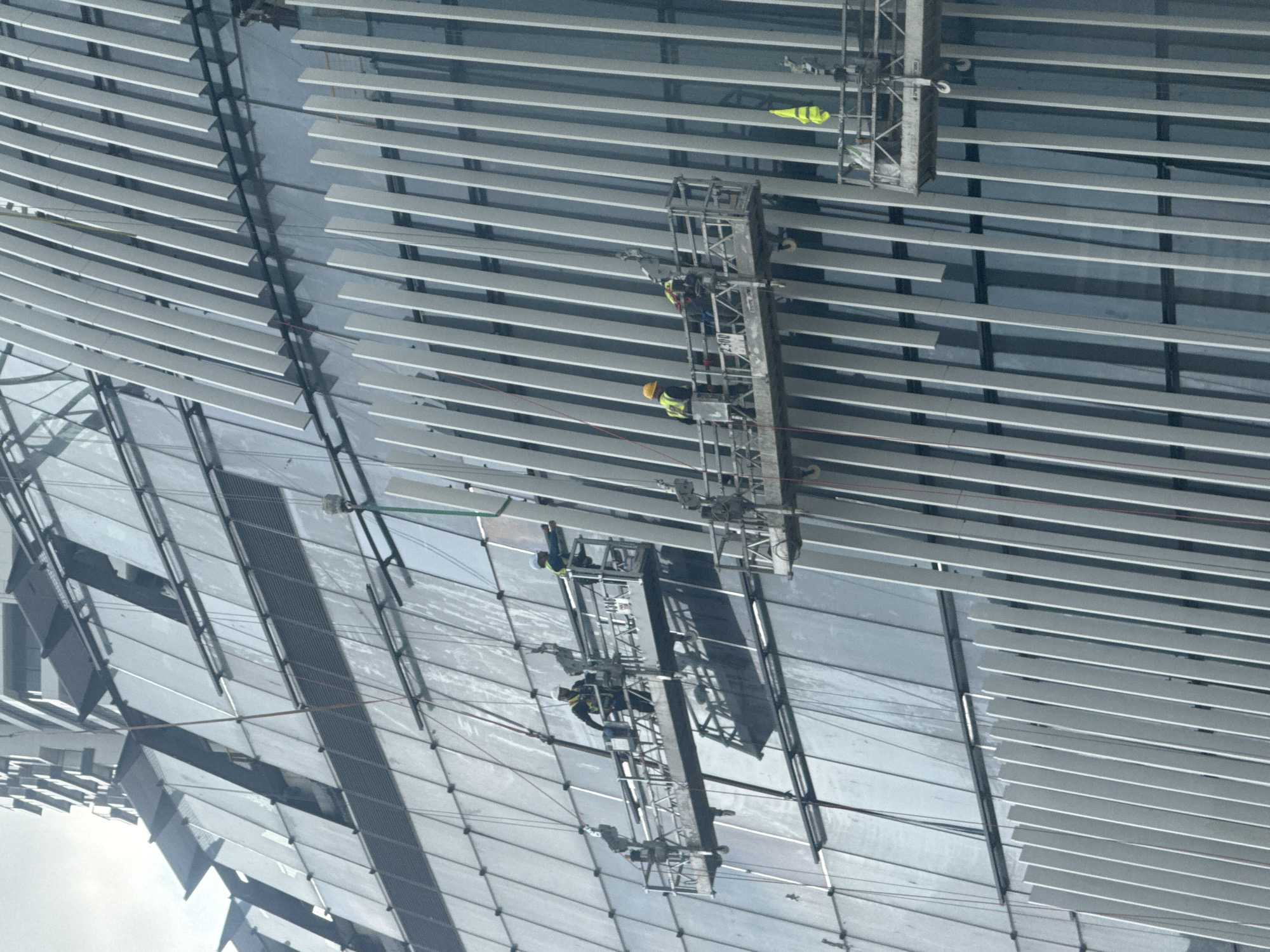 The image shows the underside of a large, curved structure, likely a stadium roof or similar architectural element in Tel Aviv, Israel. The structure is composed of parallel, horizontal slats or louvers, casting shadows and creating a textured pattern. Two maintenance or construction platforms are suspended from the structure. One platform features a worker wearing a yellow hard hat and safety vest. A yellow safety vest is attached to the upper platform as well. The platforms are positioned at different heights, suggesting ongoing work or maintenance. The sky is visible in the lower left corner, with a white gradient indicating cloud cover.