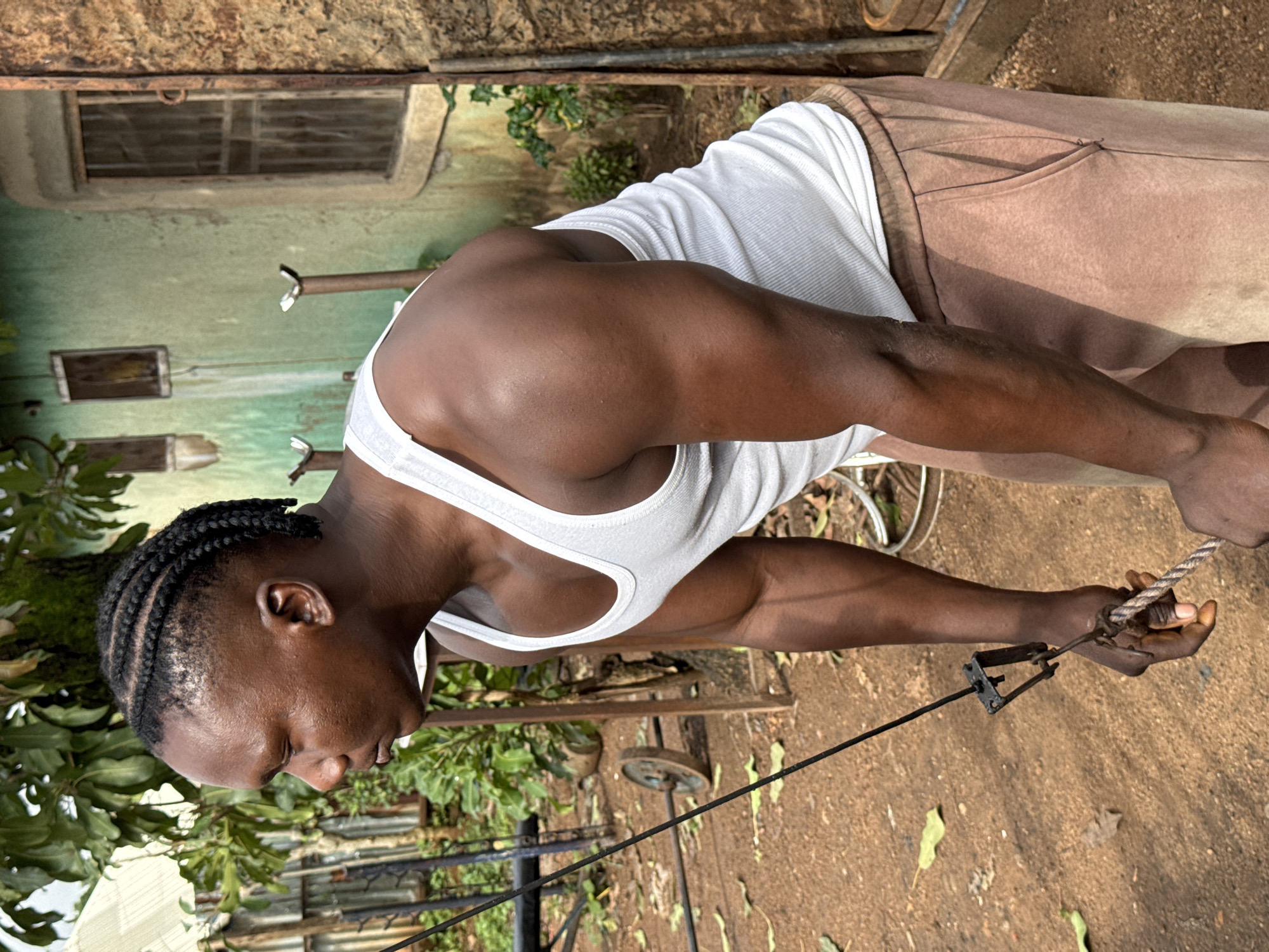 The image shows a young Black man with braided hair, wearing a white tank top and brown sweatpants. He is outdoors, with a weathered building in the background. The building appears to be made of concrete and has visible pipes and a couple of windows. The ground is dirt and covered with fallen leaves. The man is engaged in what appears to be a physical exercise, as he is pulling on a rope attached to a metal apparatus. His muscles are visible, suggesting he is working out. The lighting suggests it is daytime, possibly late morning or early afternoon, with no indication of specific weather conditions. The overall scene has a rustic and somewhat unkempt feel, consistent with the provided location context of Akwanga, Nigeria. There is no visible text in the image.