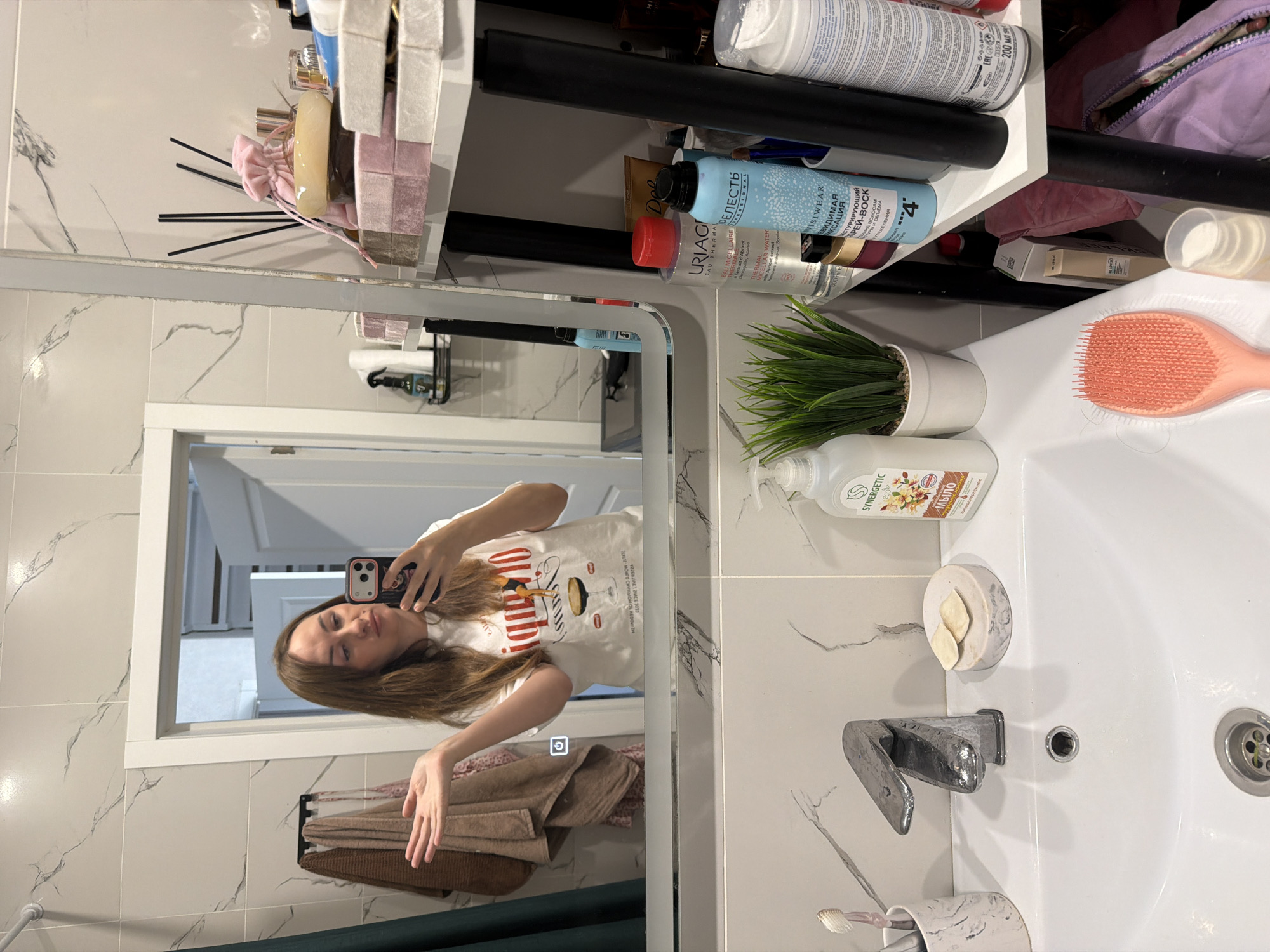 A woman is taking a selfie in a bathroom in Malinovka, Russia. She is standing in front of a mirror, holding her phone in her right hand. She is wearing a white t-shirt with a graphic print. Her left arm is outstretched, with her palm facing upwards. The bathroom features white marble tiles on the walls and a white sink with a chrome faucet. On the shelves next to the mirror, various beauty products and toiletries are visible, including bottles of shampoo, conditioner, and sprays, as well as some decorative items. A pink hairbrush rests on the edge of the sink. A rack with towels is hanging on the wall to the left of the mirror. The overall impression is of a well-stocked, modern bathroom.