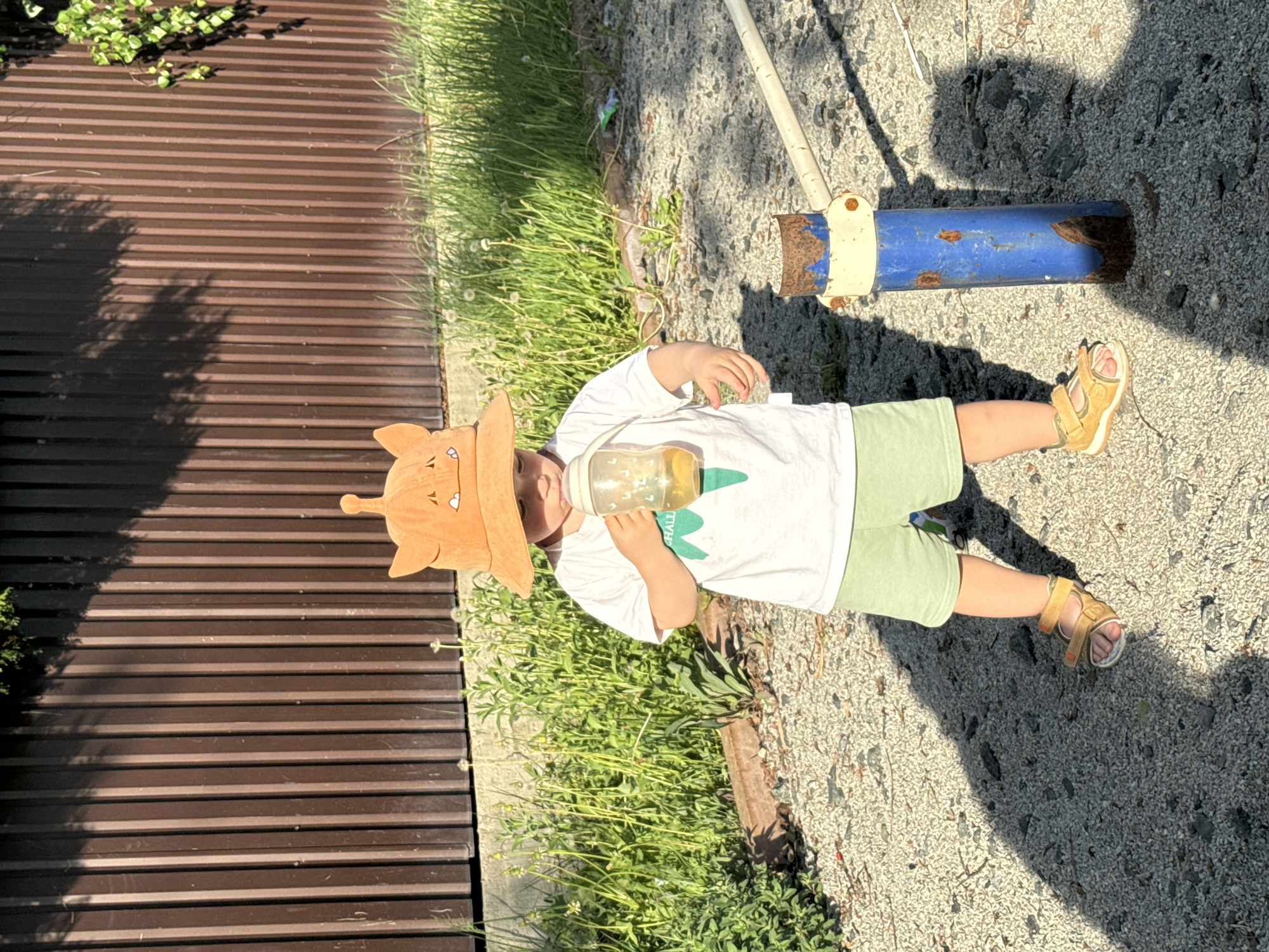 A small child wearing a brown hat with protrusions, a white t-shirt with a green print, and light green shorts is lying on their side on a gravel surface. The child is drinking from a clear bottle. The child is also wearing yellow sandals. To the child's left is a brown corrugated fence and green vegetation. Behind the child is a rusty blue metal pipe. The scene appears to be outdoors in daylight. The location is Селекционер-2 Снт, Russia.