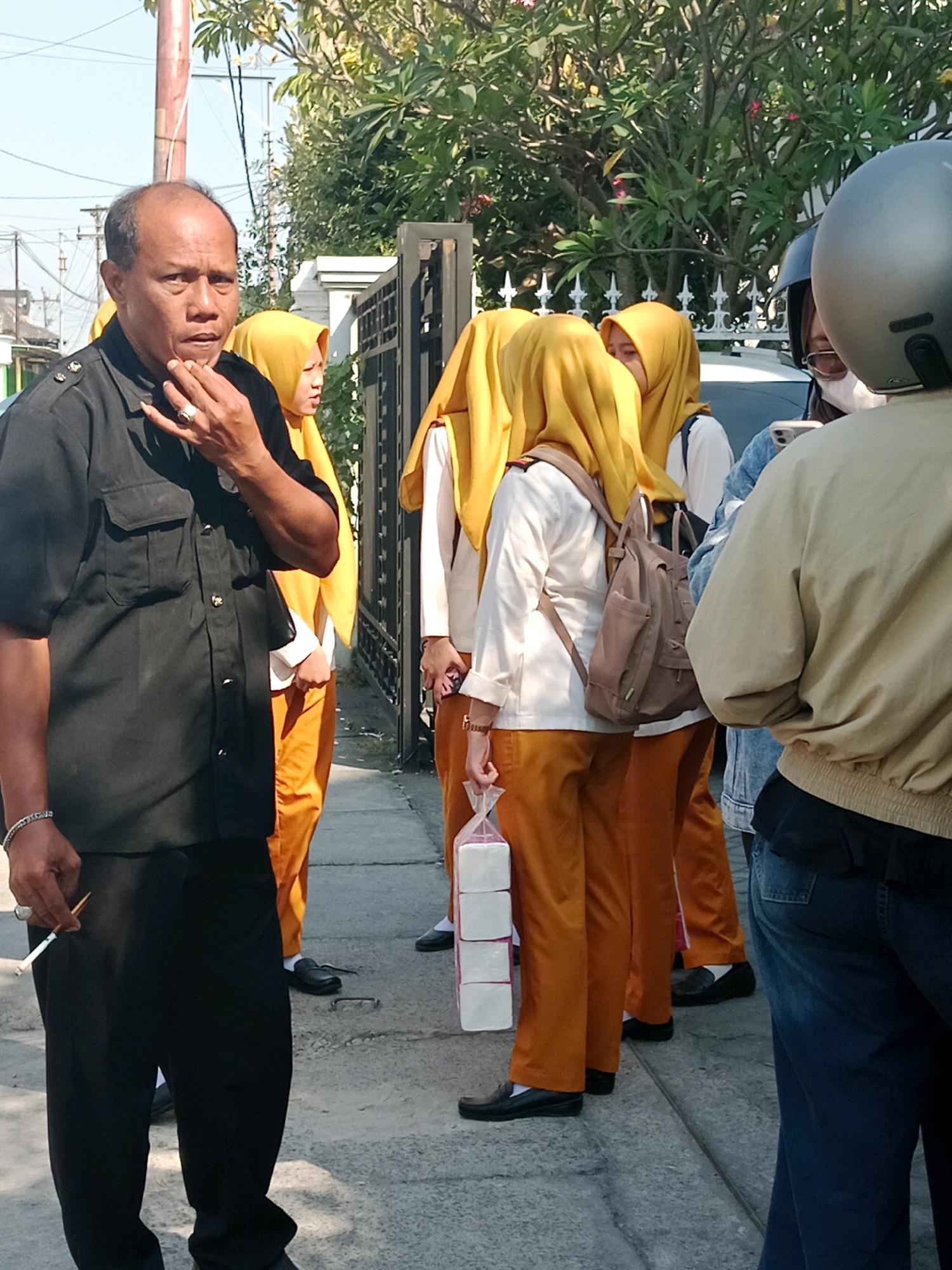 In Cemani, Indonesia, on a sunny day, a group of people are gathered outdoors on a paved walkway next to a metal gate and lush green foliage. On the left, a man with dark skin and receding hairline, wearing a black collared shirt and black pants, holds a lit cigarette in his right hand near his mouth, with a silver ring on his ring finger and a silver bracelet on his left wrist. He is looking towards the right. To his right, a young person wearing a yellow hijab and an orange uniform outfit, stands partially obscured by the man. In the center and background, several other individuals, all wearing yellow hijabs and orange uniform outfits, are visible. One of them is carrying a brown backpack. To the far right, a person wearing a light beige jacket and blue jeans is partially visible from the back, with a motorcycle helmet obscuring their head. Another person in a denim jacket and white surgical mask is also visible in the background. A stack of white packaged items, possibly tissue paper, is held by one of the individuals in orange. The ground is paved with grey concrete slabs.