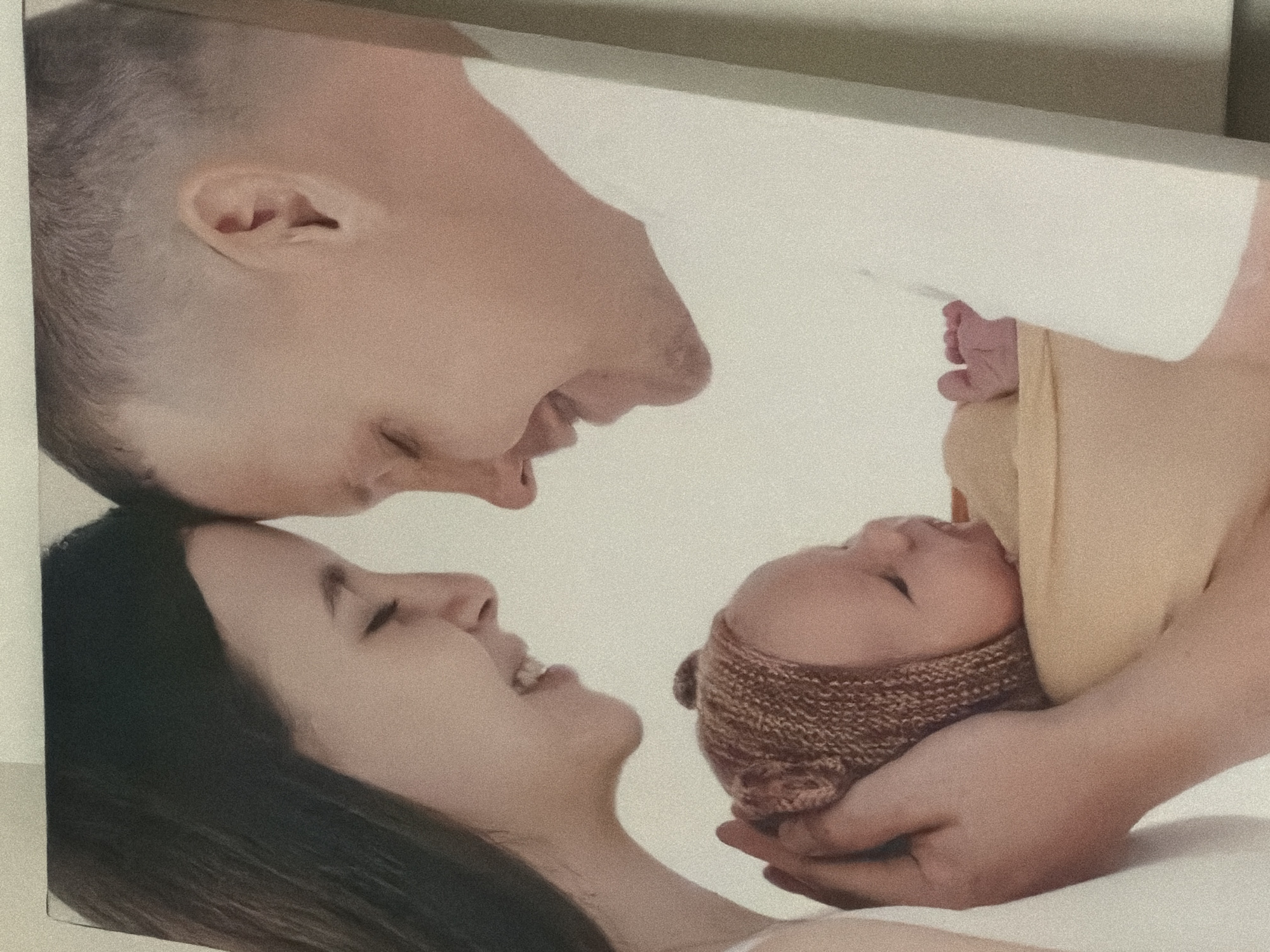 The image depicts a family portrait of a mother, father, and newborn baby. The location in the city of Chaikovsky, Russia is unknown.
The father, with a shaved head, is positioned at the top of the frame, looking down towards the baby. The mother, with long dark hair, is at the bottom, also looking towards the baby. The newborn is held in someone's hands, possibly the father's, and is wearing a knitted hat. The scene appears to be indoors with a plain white background.