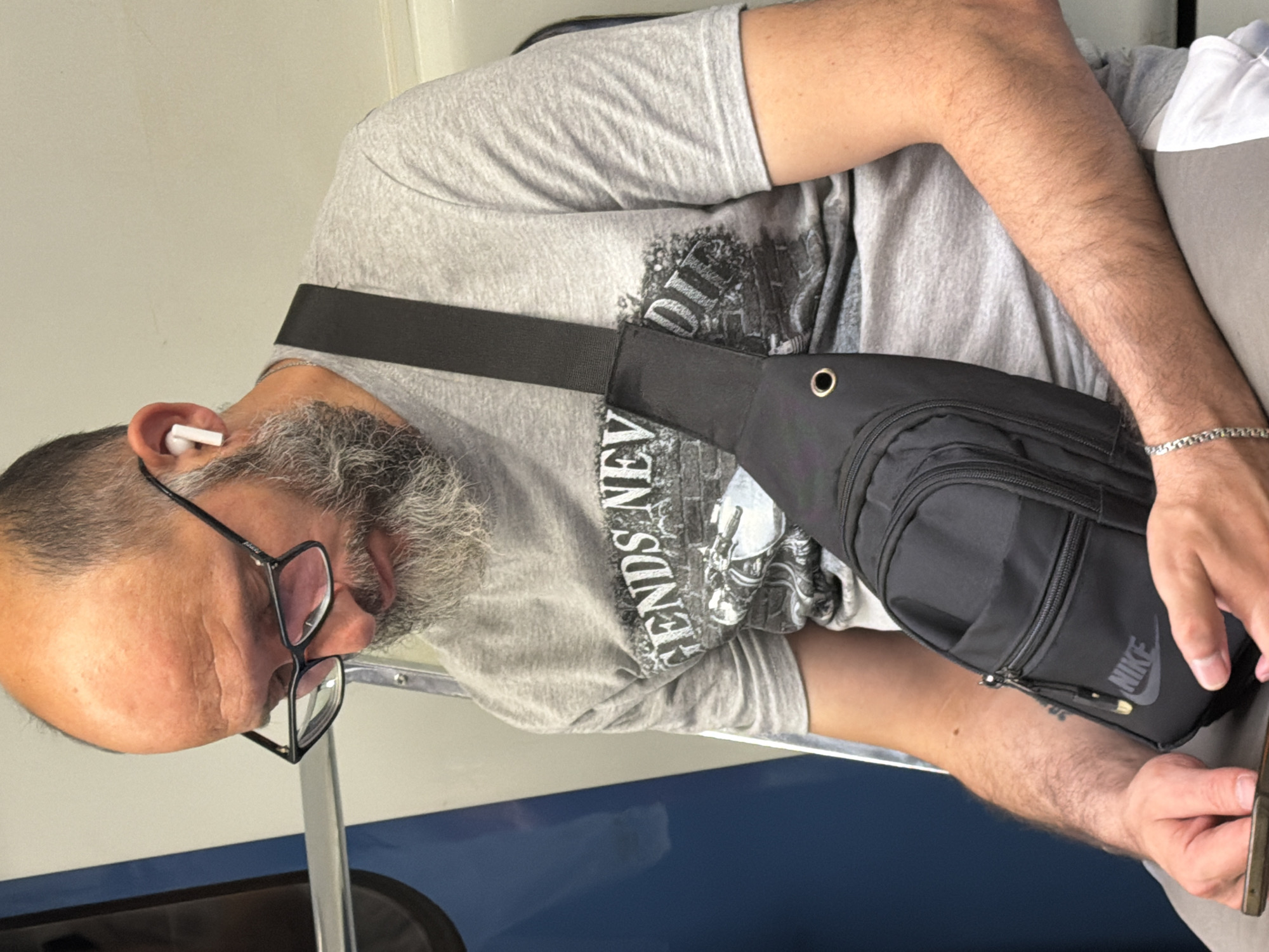 A man with a bald head, glasses, and a gray beard sits on what appears to be a train or bus, looking down at his phone. He is wearing a gray t-shirt with a graphic print and a black sling bag across his chest. White earbuds are in his ears. The environment is a clean, indoor public transportation setting with blue and white paneling. The man is holding a smartphone in his hands.