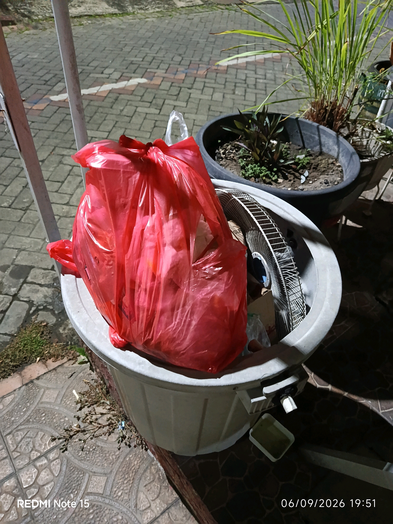 A red plastic trash bag is placed inside a large, light grey plastic bin. The trash bag is opaque and appears to contain various discarded items. Partially visible inside the bin, behind the trash bag, is the metal grill and frame of an electric fan. The bin is situated on a paved area with a patterned tile surface. In the background, there are two dark-colored plant pots. One contains a tall, leafy green plant, and the other holds smaller, dark-leaved plants. The scene is outdoors and appears to be at night or in low light conditions, as indicated by the artificial lighting and shadows. The bottom left corner of the image shows the text "REDMI Note 15", and the bottom right corner displays the date and time "06/09/2026 19:51". The location is identified as Keboananom, Indonesia.