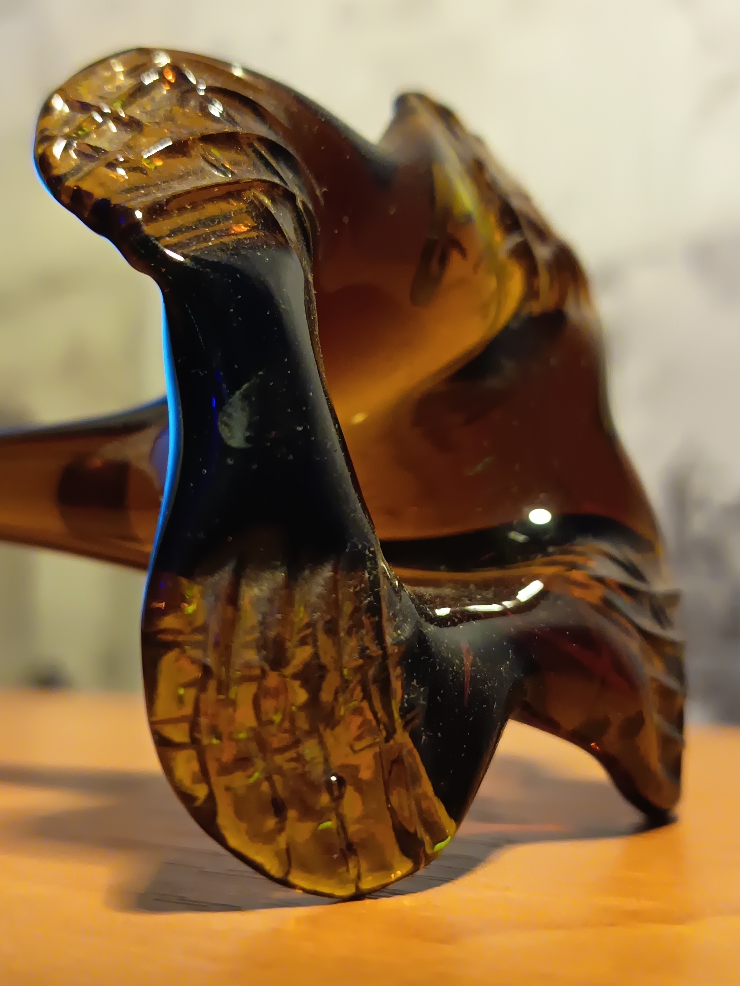The image is a close-up, low-angle shot of a dark amber glass figurine, possibly depicting a bird or mythical creature, resting on a light brown wooden surface. The figurine has intricate carvings and reflects light, creating highlights and showing internal patterns. The background is blurred and appears to be a wall with a light, textured pattern. The scene is indoors, with a light source illuminating the object from the front and slightly from the left, casting a shadow on the surface. There are no people or actions visible in the image, and no specific location in Izmail, Ukraine, can be identified.