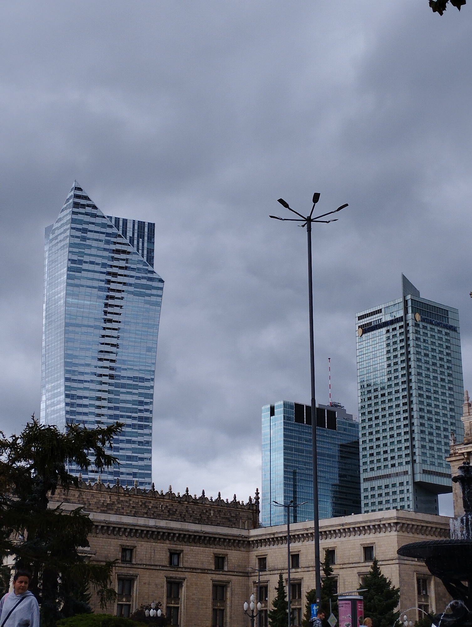 The image depicts a street-level view of Warsaw, Poland, showcasing a mix of historic and modern architecture under a cloudy sky. In the foreground, a woman in a light-colored hooded sweatshirt is partially visible on the left, looking towards the camera. Behind her, a large, dark green coniferous tree partially obscures a historic, light-brown stone building with numerous windows and ornate architectural details along its roofline. A lamppost with multiple lights stands prominently in the middle ground.
Dominating the skyline are several modern skyscrapers. The most striking is a tall, triangular-shaped building with a reflective glass facade, its geometric form angled upwards. To its right, a taller, rectangular skyscraper with a grid-like pattern of windows and blue-tinted glass is visible, bearing the "INTERCONTINENTAL" logo. Another modern building with a blue glass facade is situated to the left of the Intercontinental, featuring a distinct cut-out section. The overall atmosphere is somber due to the overcast conditions. The scene appears to be a public square or plaza, likely a well-trafficked area of the city given the presence of pedestrians and the scale of the buildings.