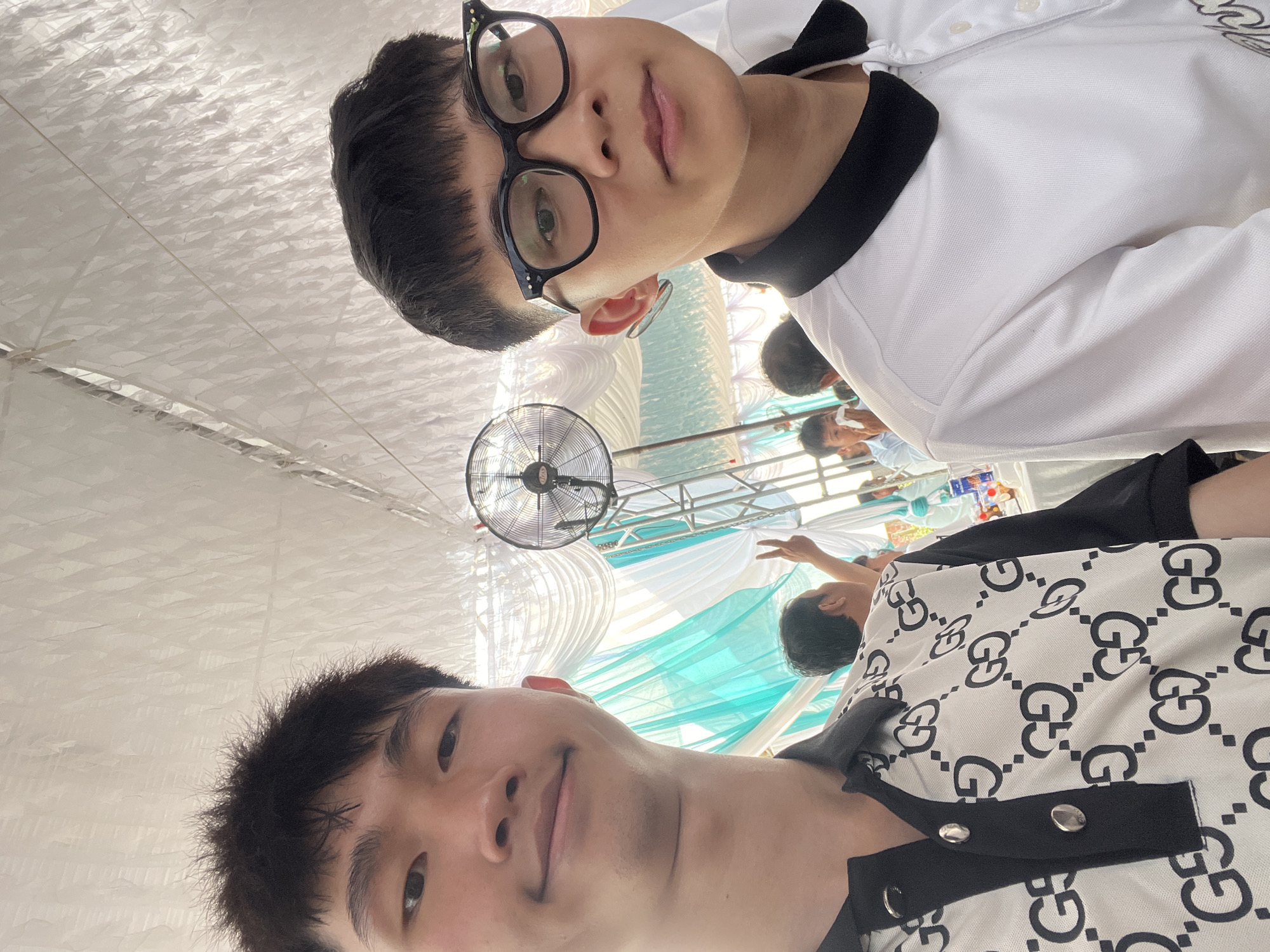 This image is a selfie featuring two young men at what appears to be a social gathering or event in Bình An, Vietnam. The photo is taken from a low angle, looking upwards.
In the foreground, the top subject is a young man with short dark hair, wearing black-rimmed glasses and a white polo shirt with black trim. He looks directly at the camera with a neutral expression. Below him, the second young man, also with short dark hair, smiles slightly while looking into the camera. He wears a white short-sleeved shirt patterned with a black interlocking 'G' logo.
The background reveals an event space adorned with white and light blue/teal fabric drapes. A black industrial-style fan is visible, hanging in the mid-ground on the left. The ceiling is covered with white pleated fabric, suggesting a temporary structure or marquee. Several other individuals are present in the background, out of focus; one person on the right is seen making a peace sign. On a table further back, small packaged items, possibly food or drinks, are visible. The bright lighting suggests it is daytime. The presence of the fan implies warm weather conditions. The overall atmosphere appears casual and friendly.