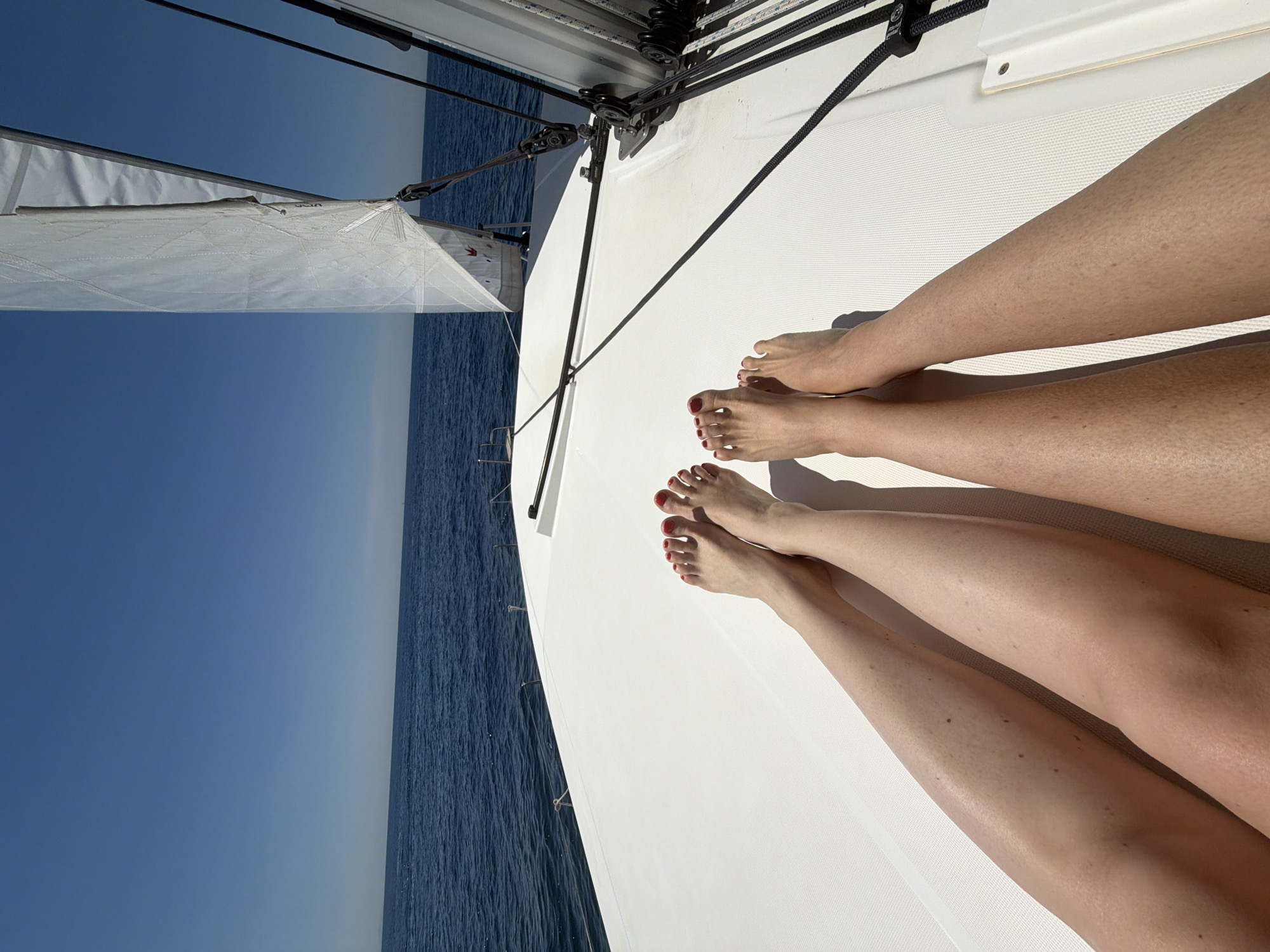 The image, taken from a low angle, depicts a scene on a sailboat at sea, possibly near Conca dei Marini, Italy, based on the presence of an ocean horizon. Two pairs of human legs and feet are visible in the foreground, positioned on the white deck of the boat. The feet in the bottom pair have red nail polish. The sailboat's rigging and a portion of its sail can be seen at the top and left side of the image. The ocean and sky fill the left side of the frame, exhibiting a clear horizon line. The water appears calm with a deep blue color, while the sky is a lighter, clear blue. The lighting suggests a sunny day. The image is rotated counter-clockwise approximately 90 degrees.