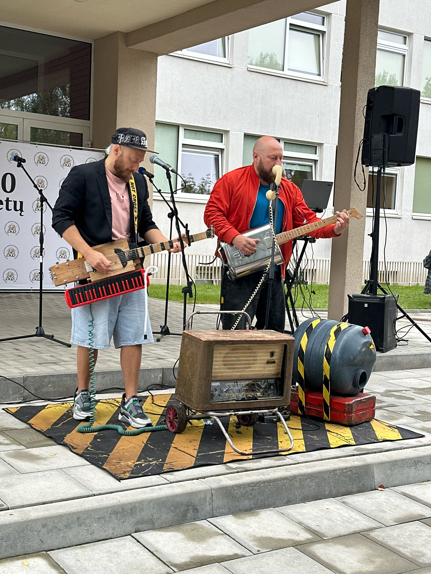Two men are playing musical instruments outdoors in front of a building. The man on the left wears a black jacket over a pink t-shirt and denim shorts, and plays a unique guitar with a keyboard attachment. He has a beard and is wearing a black baseball cap. The man on the right wears a red jacket over a blue shirt and dark pants, and plays a metallic guitar. He has a beard and is holding a vintage telephone receiver to his mouth. They are standing on a rug with black and yellow stripes. In the foreground, there is an old radio on wheels and a grey barrel with yellow and black warning stripes on a red base. The building behind them is modern with multiple windows. The scene appears to be set up for a street performance in Vilnius, Lithuania.