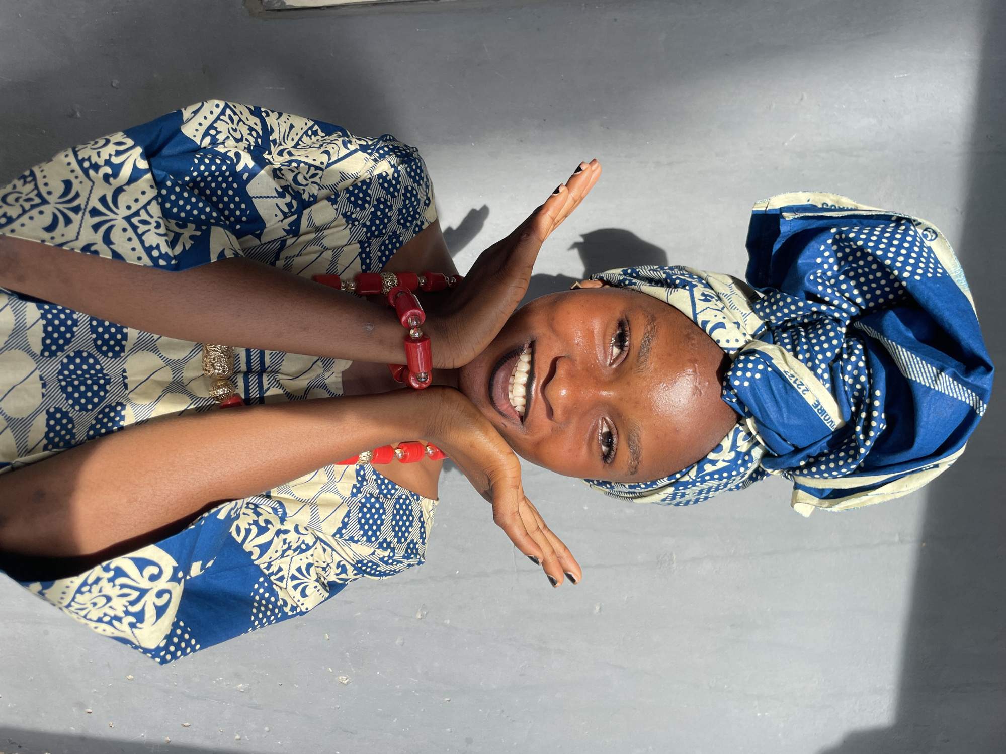 A young Black woman wearing a blue and white patterned dress and headwrap, and a red beaded necklace, is smiling and holding her hands near her face. She is likely in Badagry, Nigeria, based on the style of clothing. The background is a smooth, gray surface, possibly a floor. Sunlight creates shadows and highlights on the subject and the background.