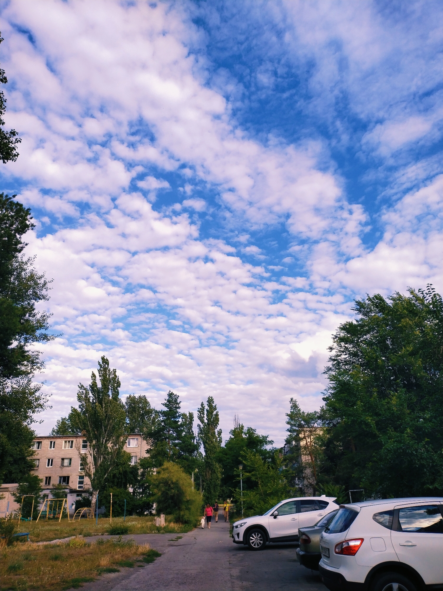 The image depicts a residential area in Horishni Plavni, Ukraine, on a day with a partly cloudy sky. In the foreground, a paved walkway leads past parked cars. Three white SUVs are visible, with one in the foreground on the right, another in the middle, and a third on the left. A darker-colored car is parked between the two white SUVs on the left. Two people are walking on the walkway, one in a pink top and the other in a yellow top, accompanied by a small white dog. In the midground, a multi-story apartment building is visible, with several windows on its facade. Adjacent to the building is a playground with swings and climbing structures. Surrounding the buildings and playground are various trees and greenery, including tall, slender trees and denser foliage. The sky above is a vibrant blue, dotted with numerous white, puffy clouds. The overall scene suggests a typical day in a residential neighborhood.