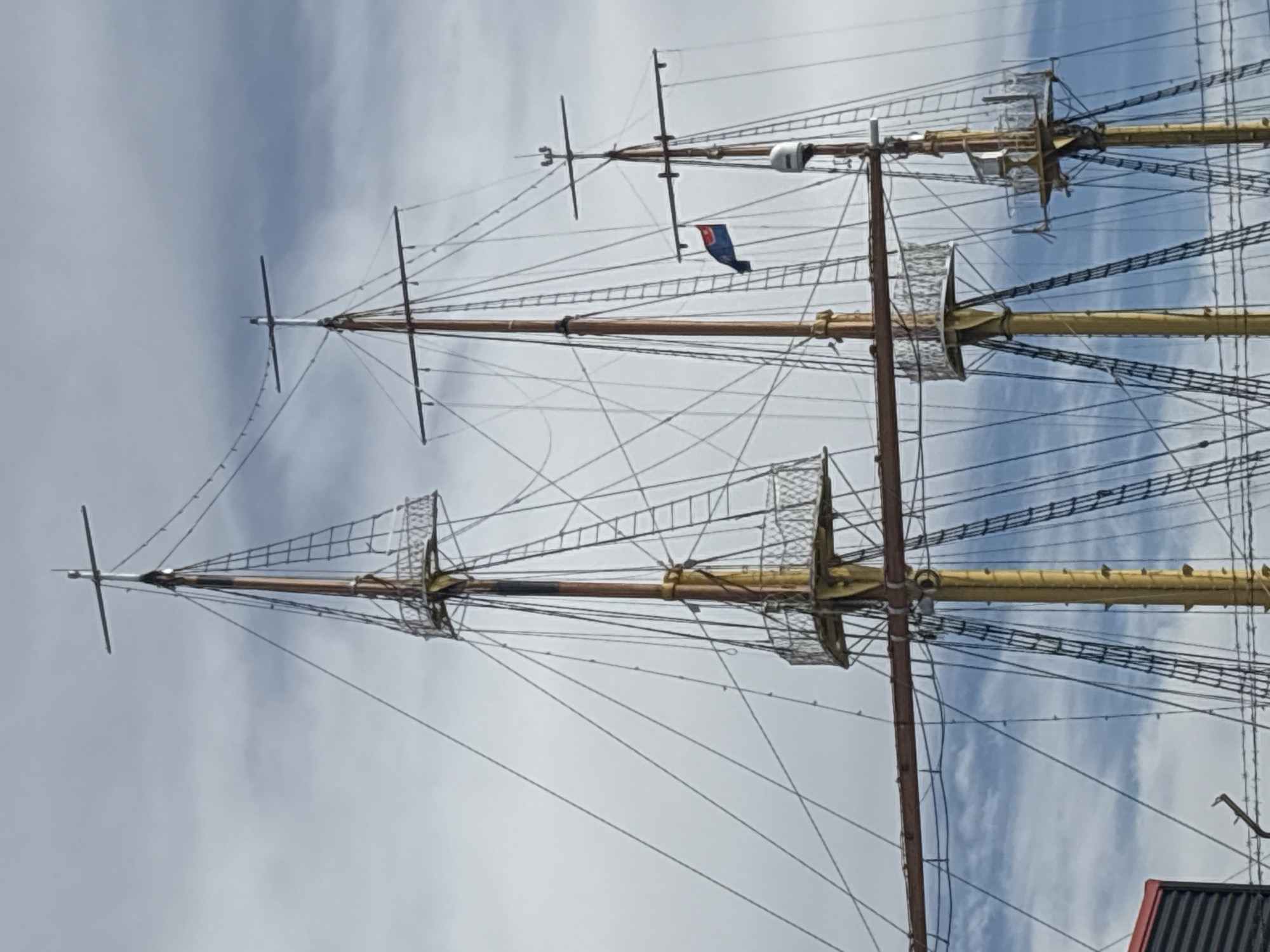 The image shows the masts and rigging of a tall ship. The masts are tall and taper toward the top, with horizontal yards extending from them. Complex network of ropes and lines, known as rigging, connect the masts and yards. A small blue and red flag is visible near the top of one of the masts. The background consists of a cloudy sky. The masts and rigging suggest that this is a ship from a historical era, likely a replica or a preserved vessel. This could be a part of the maritime museum or a tourist attraction. There is no indication in the image that the scene is in the city of Bar, Montenegro.