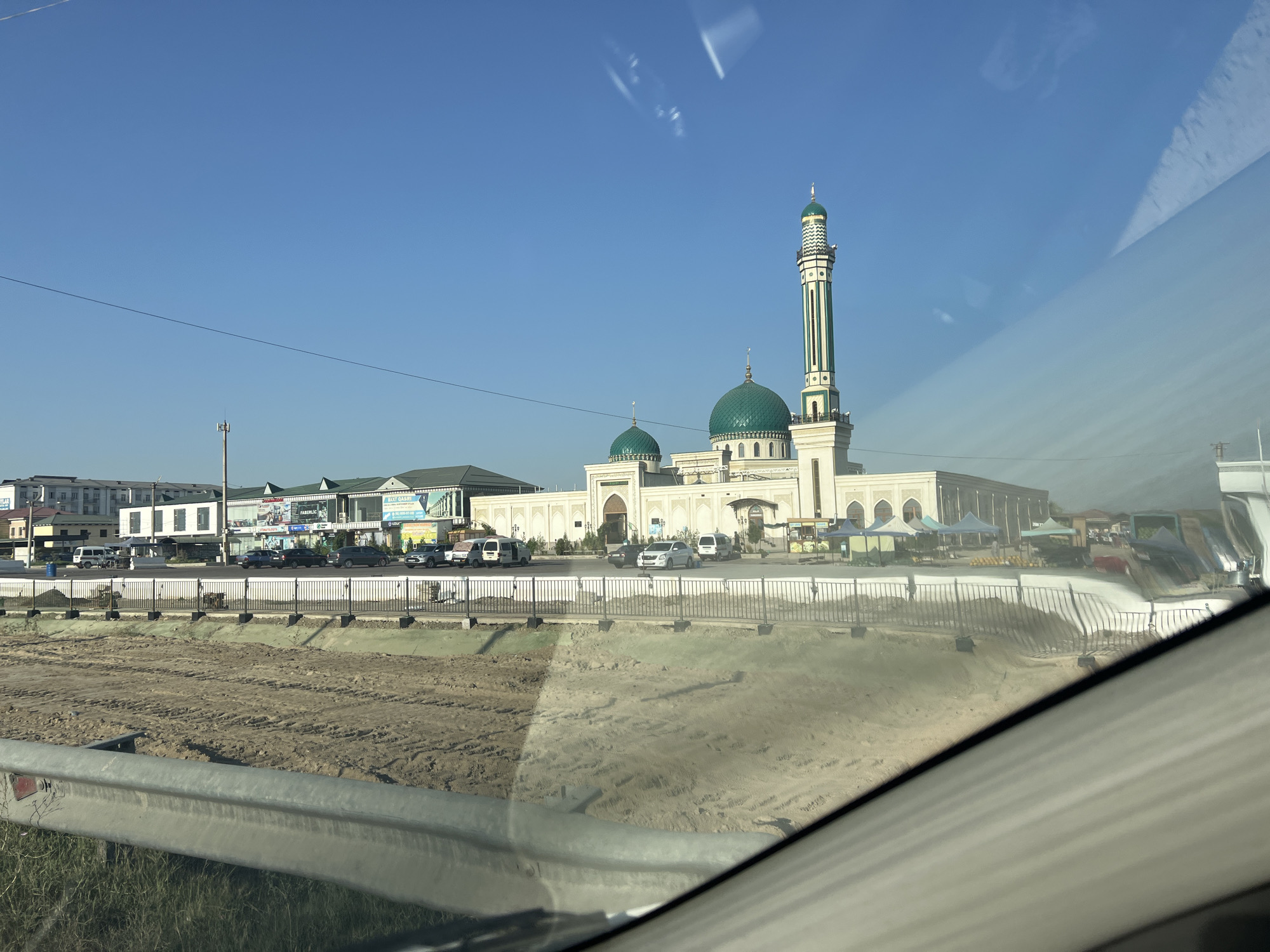 A wide-angle, eye-level shot captures a modern mosque in Baxt, Uzbekistan. The mosque features a tall, slender minaret with green and white stripes, topped with a green dome. Two smaller green domes are visible on the main building. The mosque's facade is white with decorative arches. In front of the mosque, a paved area is filled with several parked cars, including white vans and dark sedans. Beyond the mosque, a line of multi-story buildings with flat roofs stretches across the background. The scene is framed by a reflective surface, likely a car window, which casts streaks of light and reflections across the image. In the foreground, a muddy construction site with visible tire tracks is separated from the mosque area by a black metal fence. The sky is clear and blue, indicating fair weather.