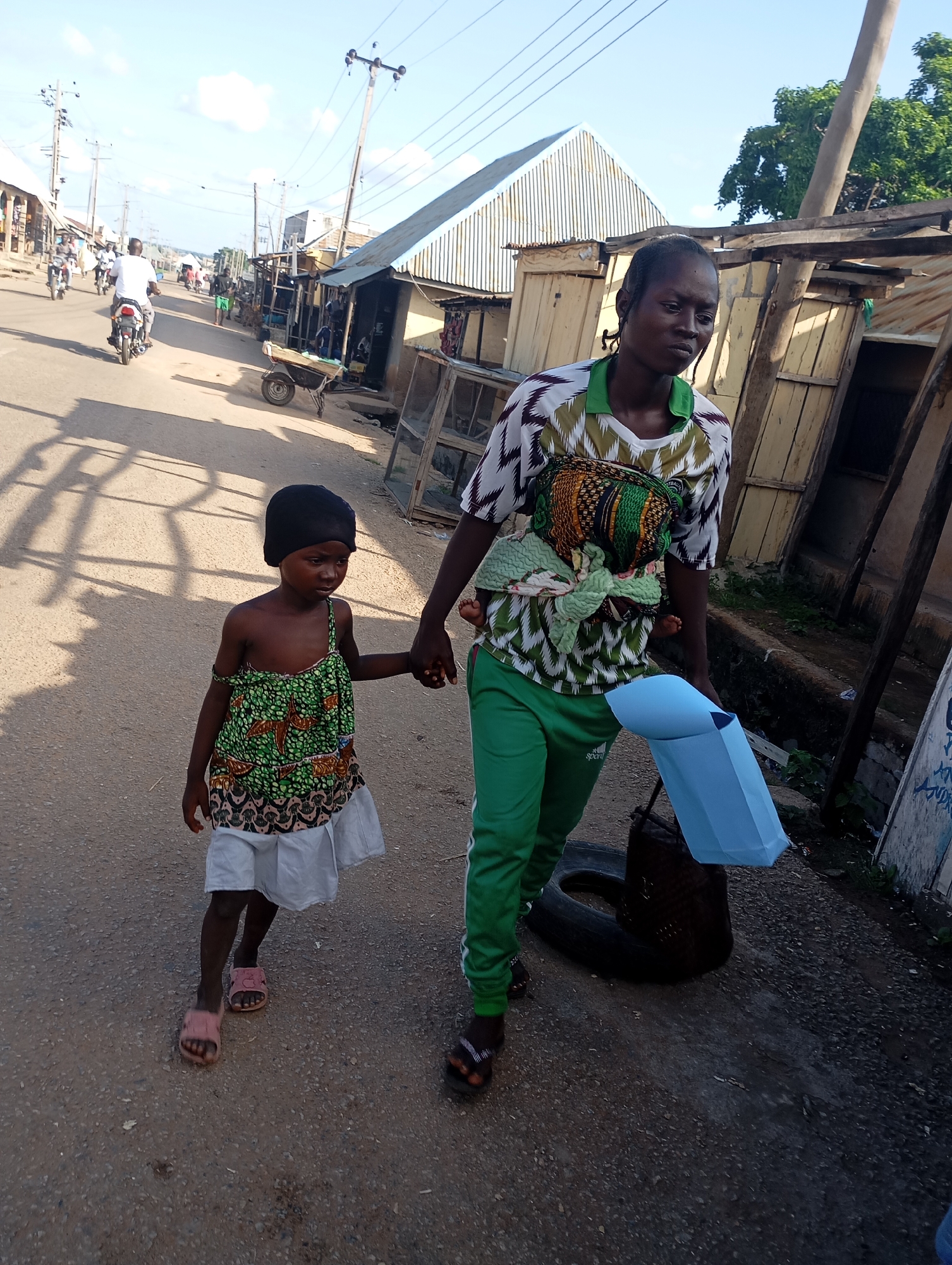 In Shendam, Nigeria, a woman, likely a mother, walks down a street carrying a baby wrapped in a cloth on her back. She is wearing a green and white patterned shirt and green track pants. She holds the hand of a young girl who is walking beside her. The girl is wearing a green and patterned sleeveless dress and pink sandals. The street is paved and lined with small buildings, some appearing to be shops or homes. There are also power lines and poles visible, typical of an urban environment. Several motorcycles are seen in the background, suggesting this is a busy thoroughfare. A car tire and a blue plastic object are on the ground near the woman. The scene depicts everyday life in the city.
