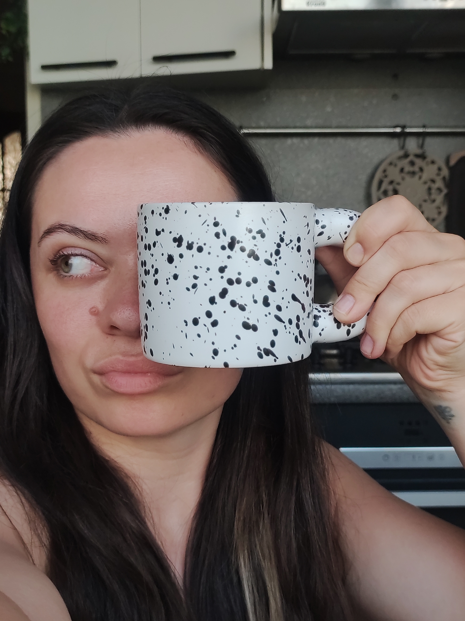 A woman with dark hair is holding a white mug with black spots in front of her face, covering her right eye. She is looking to her left with a slight pout. Her left eye is visible, and a mole is on her left cheek. She is indoors, and behind her are kitchen cabinets and a metal rod with cooking utensils hanging from it. A tattoo is visible on her right wrist. The location is not identifiable as Chaikovsky, Russia, based on the provided image.
