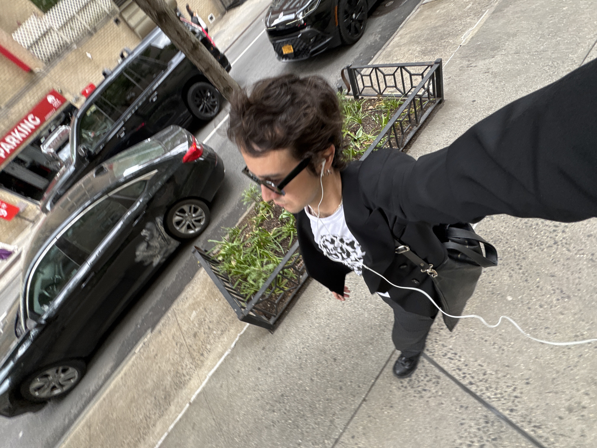 A person takes a selfie on a sidewalk in New York City. The individual wears sunglasses, earbuds, a white t-shirt with a graphic, a black blazer, gray trousers, and black shoes. A black bag hangs from their shoulder. The sidewalk has a small metal-framed planter box with greenery. Cars and SUVs are parked at the curb, and a "PARKING" sign is visible. The cars are black. The sidewalk is concrete. The background includes a brick building with windows.
