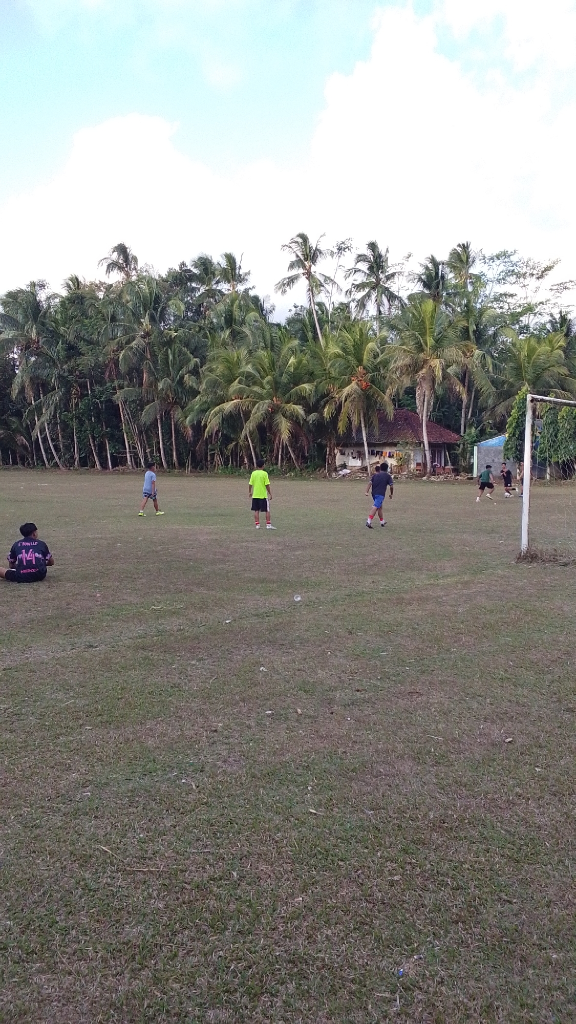The image depicts an outdoor scene on a grassy field, likely a soccer field, with a grove of palm trees and a small building in the background. The sky above is a pale blue with white clouds. In the foreground, a person is sitting on the grass, facing away from the camera. Further into the field, several individuals appear to be playing soccer. One person in a bright green shirt and black shorts is standing in the middle of the field. Two individuals in dark shirts and shorts are positioned closer to a soccer goal, with one actively kicking a ball. Another person in a light blue shirt and dark shorts is standing to the left, facing away. The environment suggests a rural or semi-rural setting in Cintaratu, Indonesia, given the prevalence of palm trees and the architectural style of the building in the background.